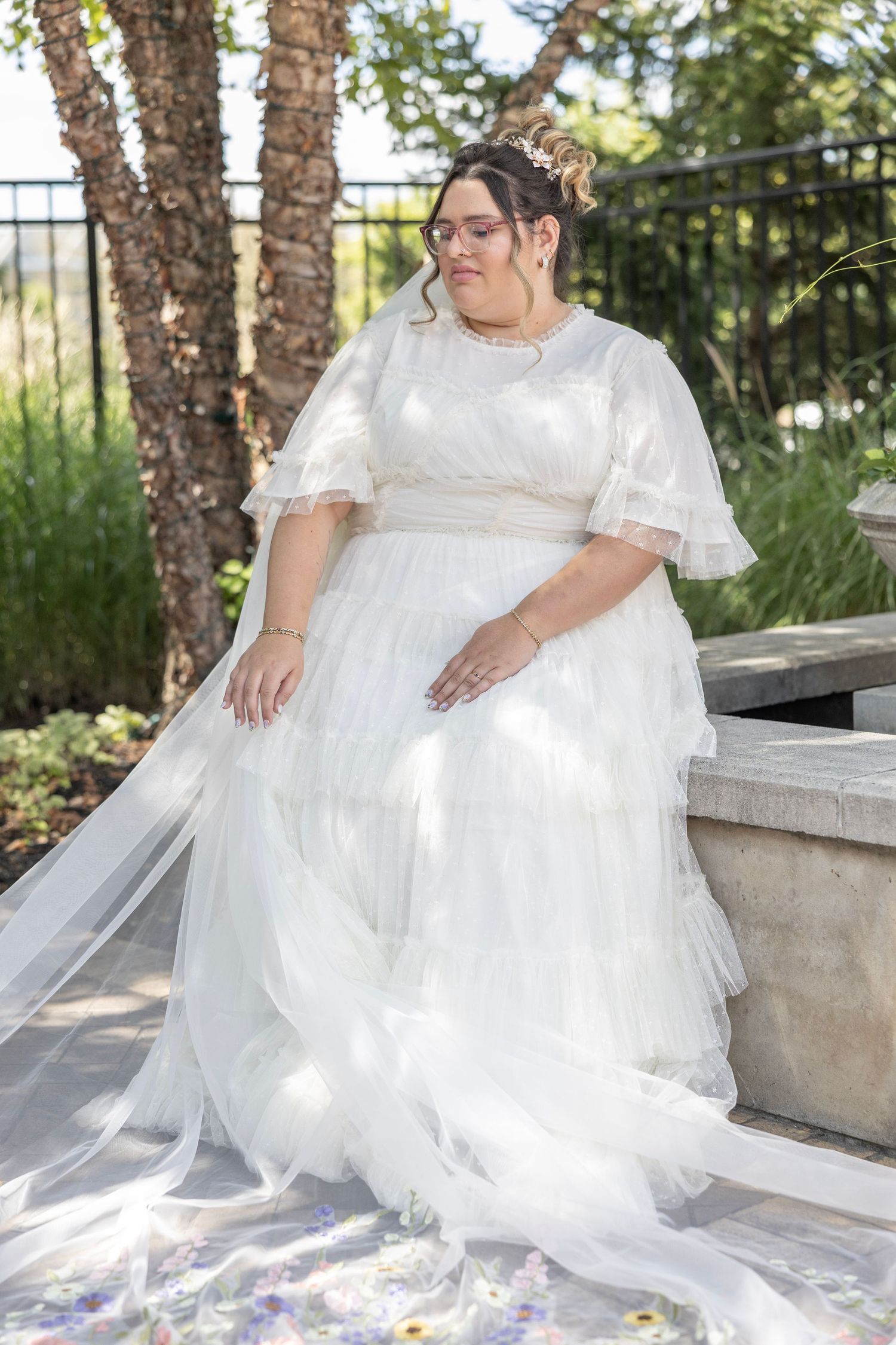 A bride in a flowing white dress sits gracefully on stone steps in a sunlit garden setting.