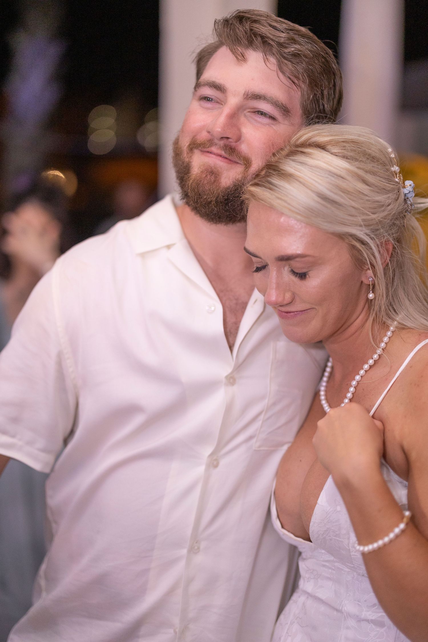 A couple shares an intimate moment at a wedding reception, with the bride in white and groom in casual attire.