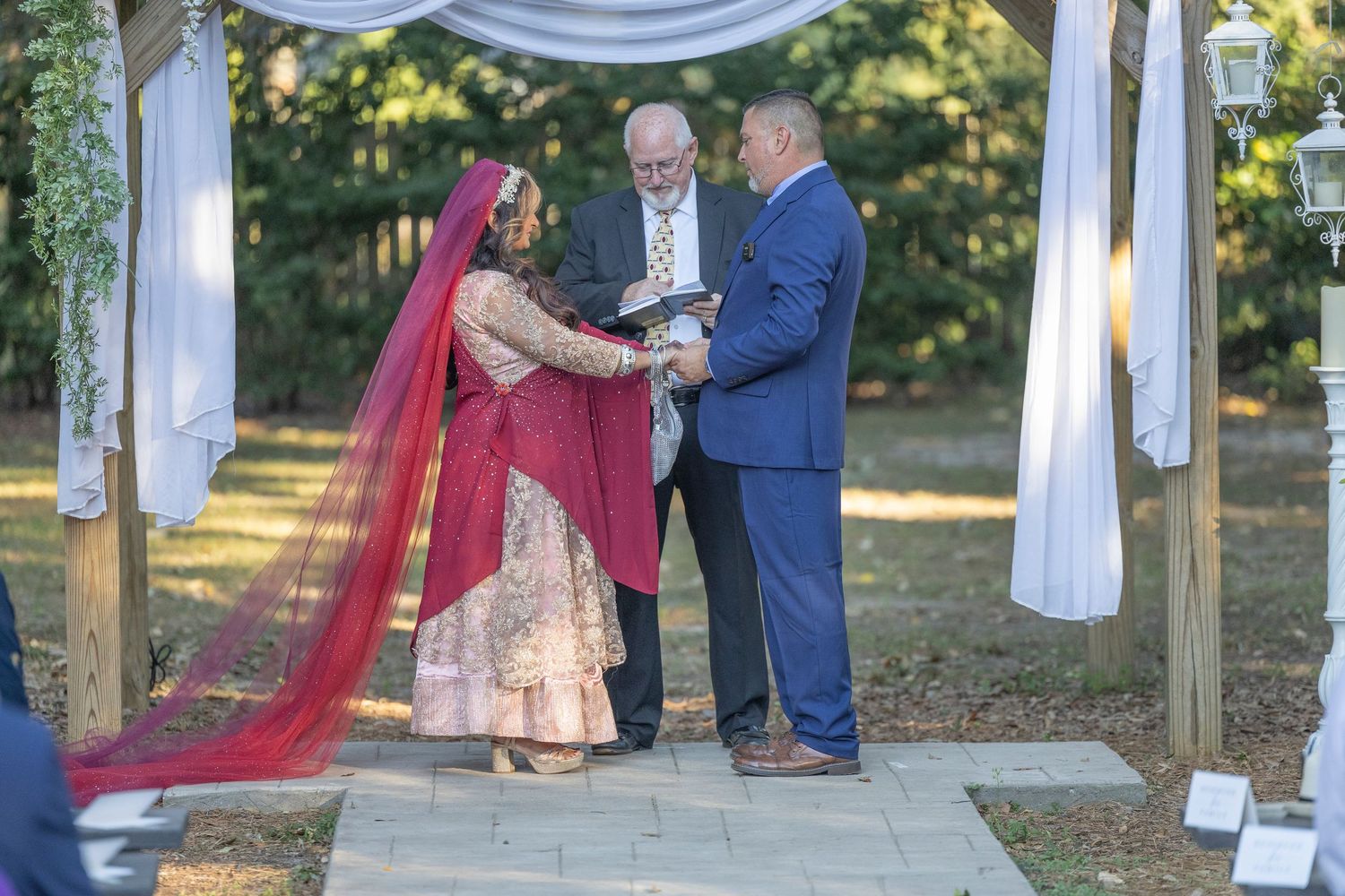 A couple exchanges vows during an outdoor wedding ceremony under a white draped gazebo in a wooded setting.