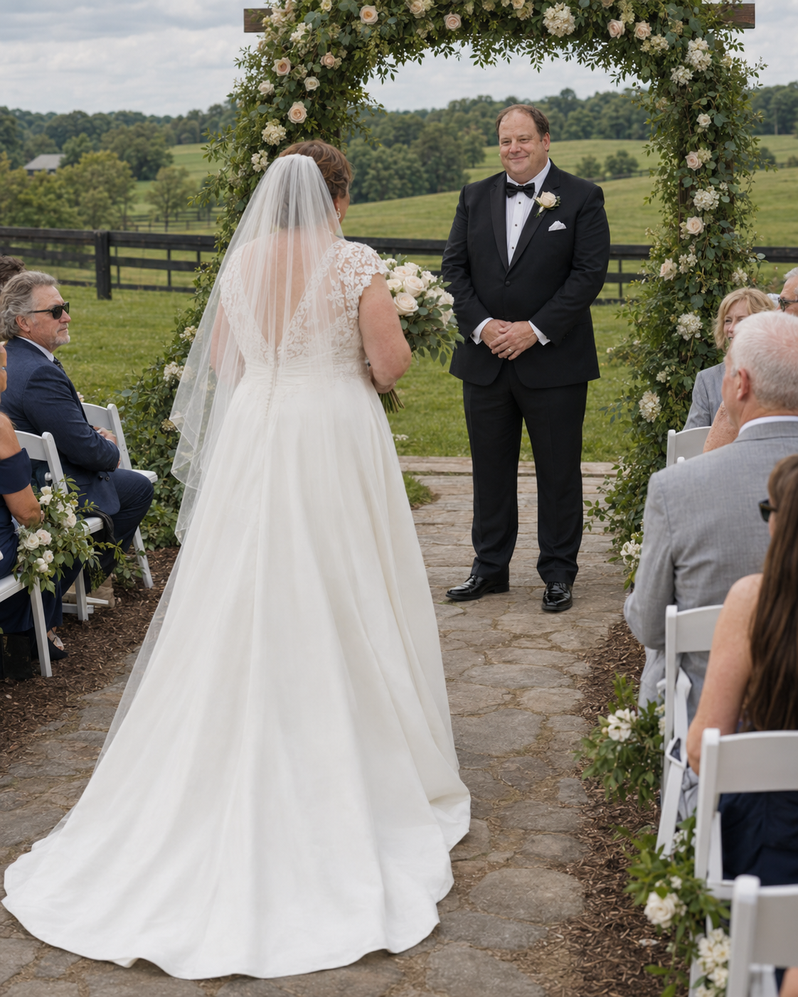 Bride walking down outdoor aisle toward smiling groom under floral arch at farm wedding.