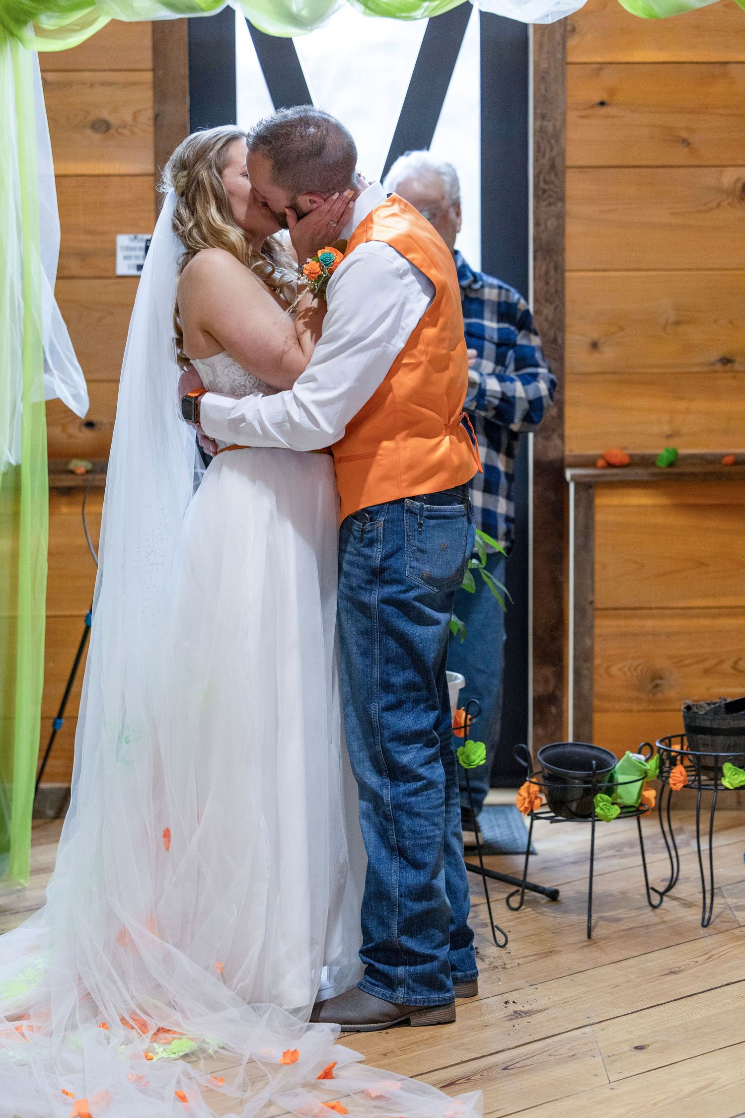 Bride and groom share their first kiss at a rustic barn wedding, with green draping and orange accents.