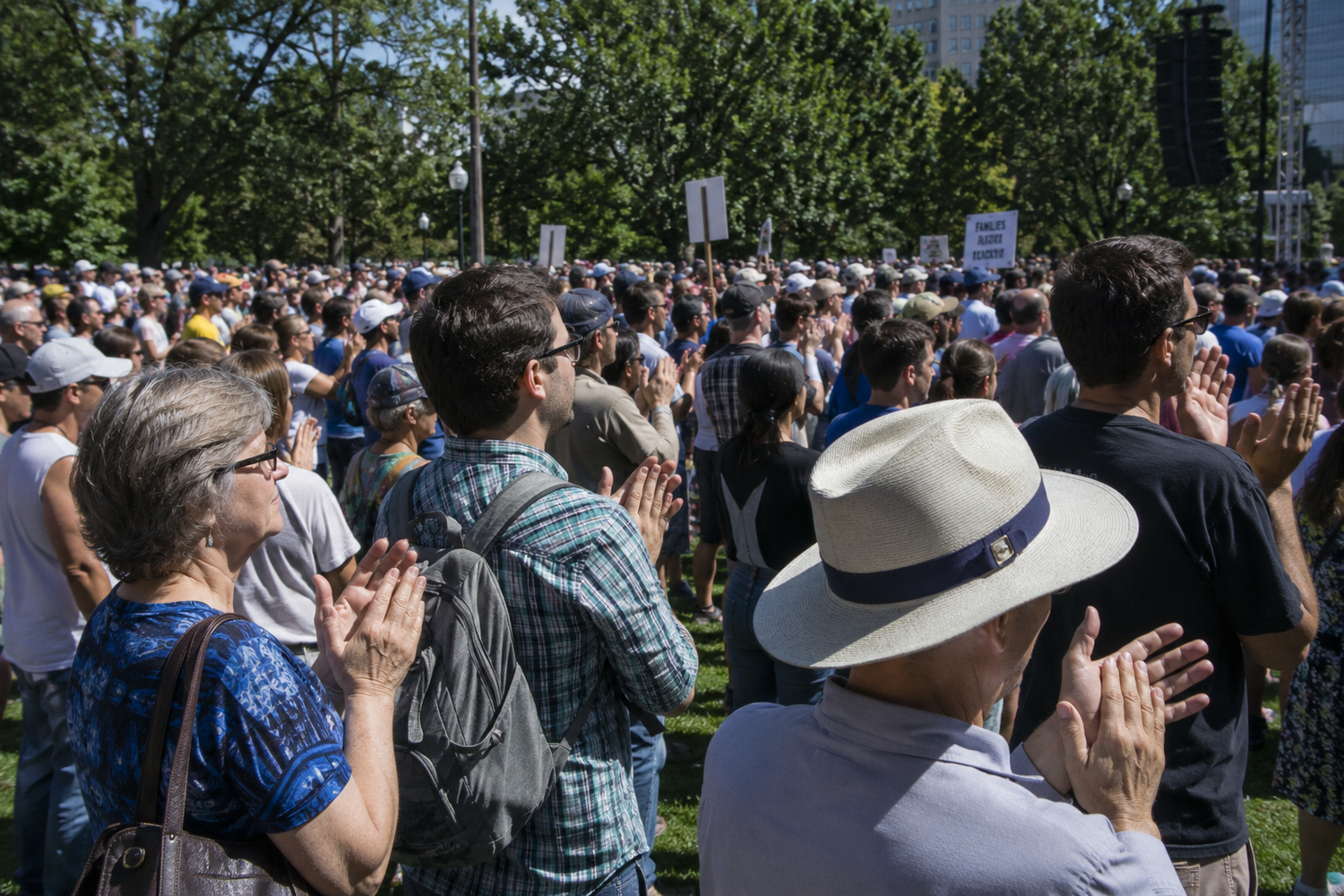 Diverse crowd applauds at outdoor public gathering in sunny park with protest signs visible.