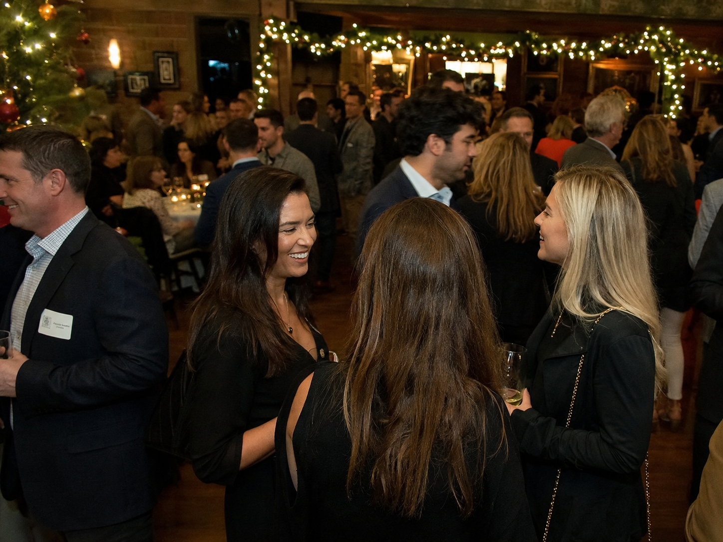 Guests mingling at an elegant holiday party with warm string lights and festive decorations in a rustic venue.