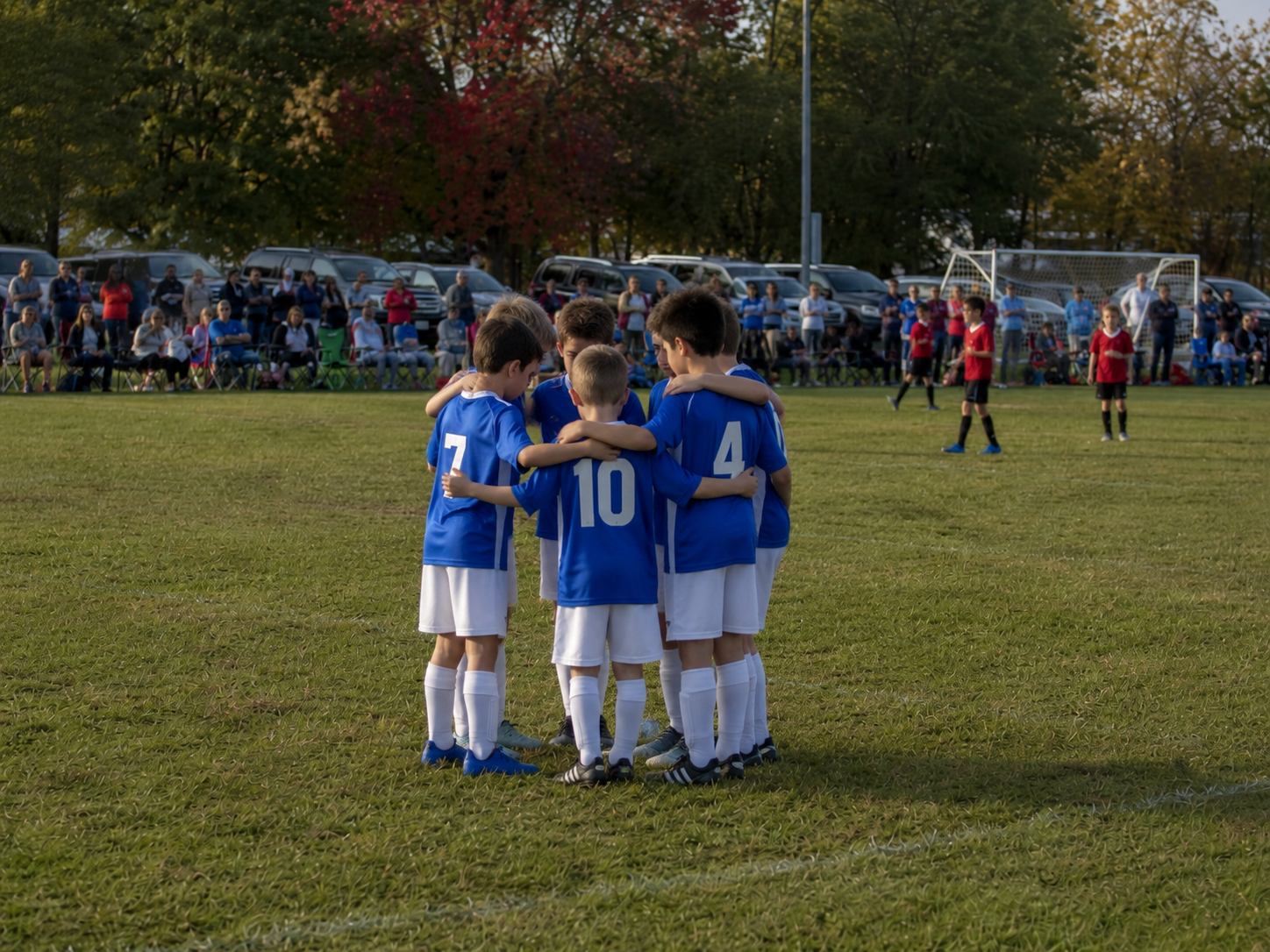 Young youth soccer players in blue uniforms huddling together on a green field during an autumn game.