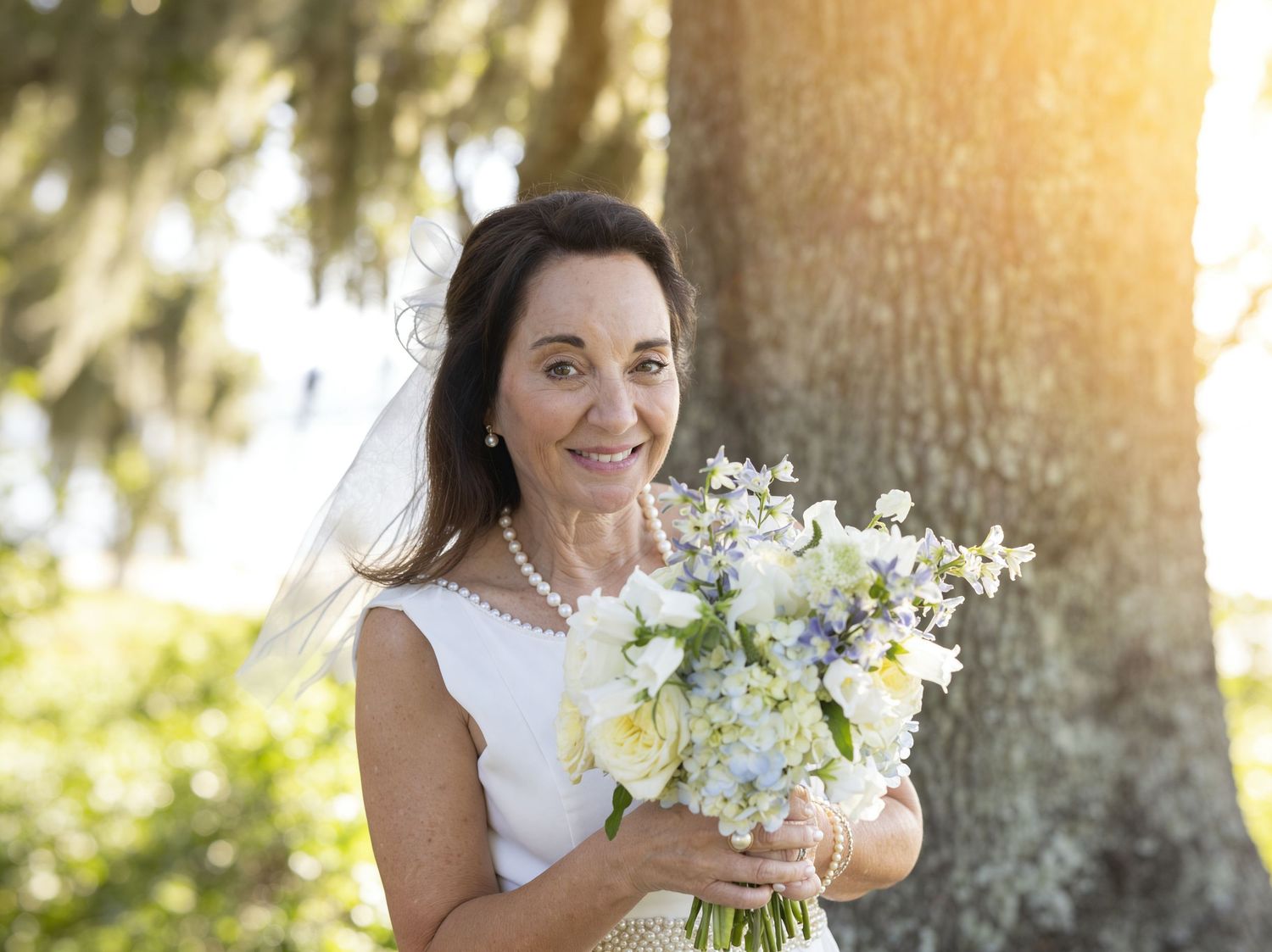 Elegant bride holding white and blue bouquet, wearing veil and pearl necklace, standing near tree in outdoor setting.