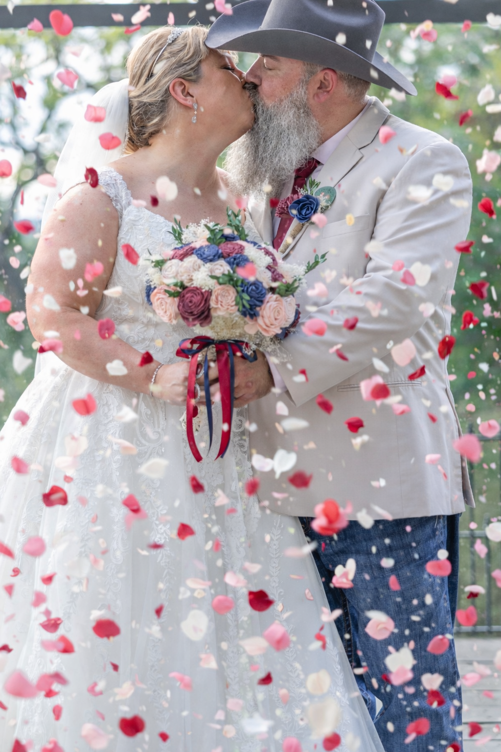 Bride and groom share a kiss surrounded by falling rose petals, bride holding a pink and blue bouquet.