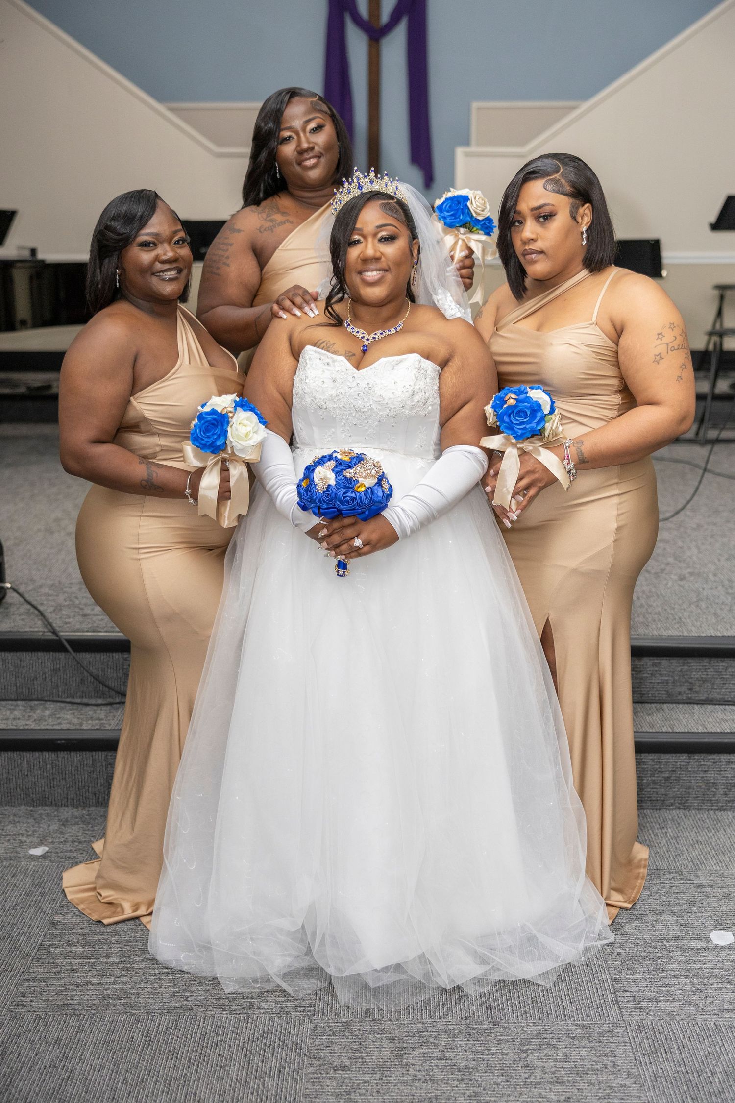 Bride in white gown with three bridesmaids in gold dresses holding blue and white bouquets inside a church.