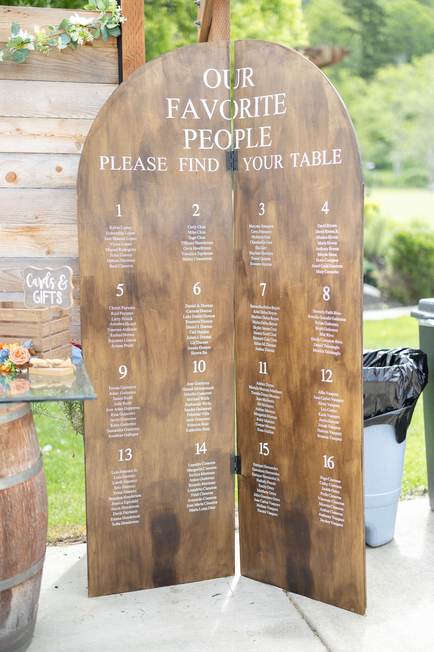 Rustic wooden seating chart displays table assignments on multiple stained panels for an outdoor wedding reception.