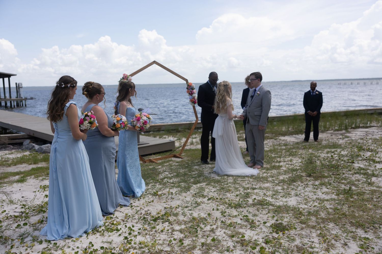 A group gathers for a seaside wedding ceremony on a rocky beach with mountains in the background.