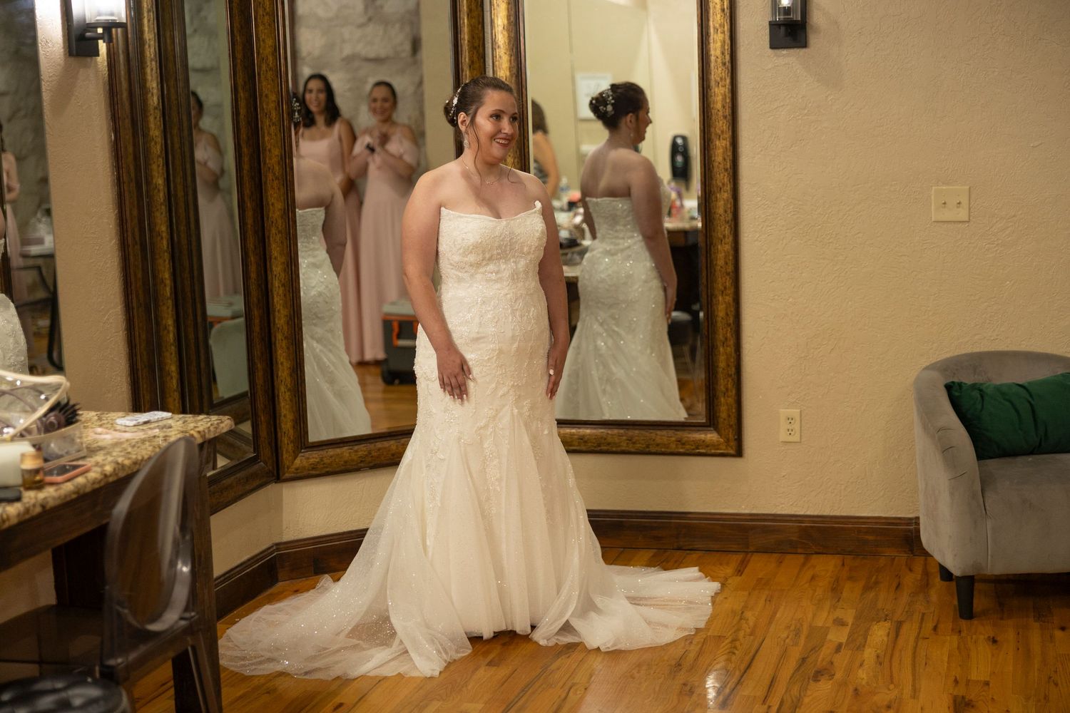 A bride in a fitted lace mermaid wedding gown stands in a bridal suite with mirrors and hardwood floors.