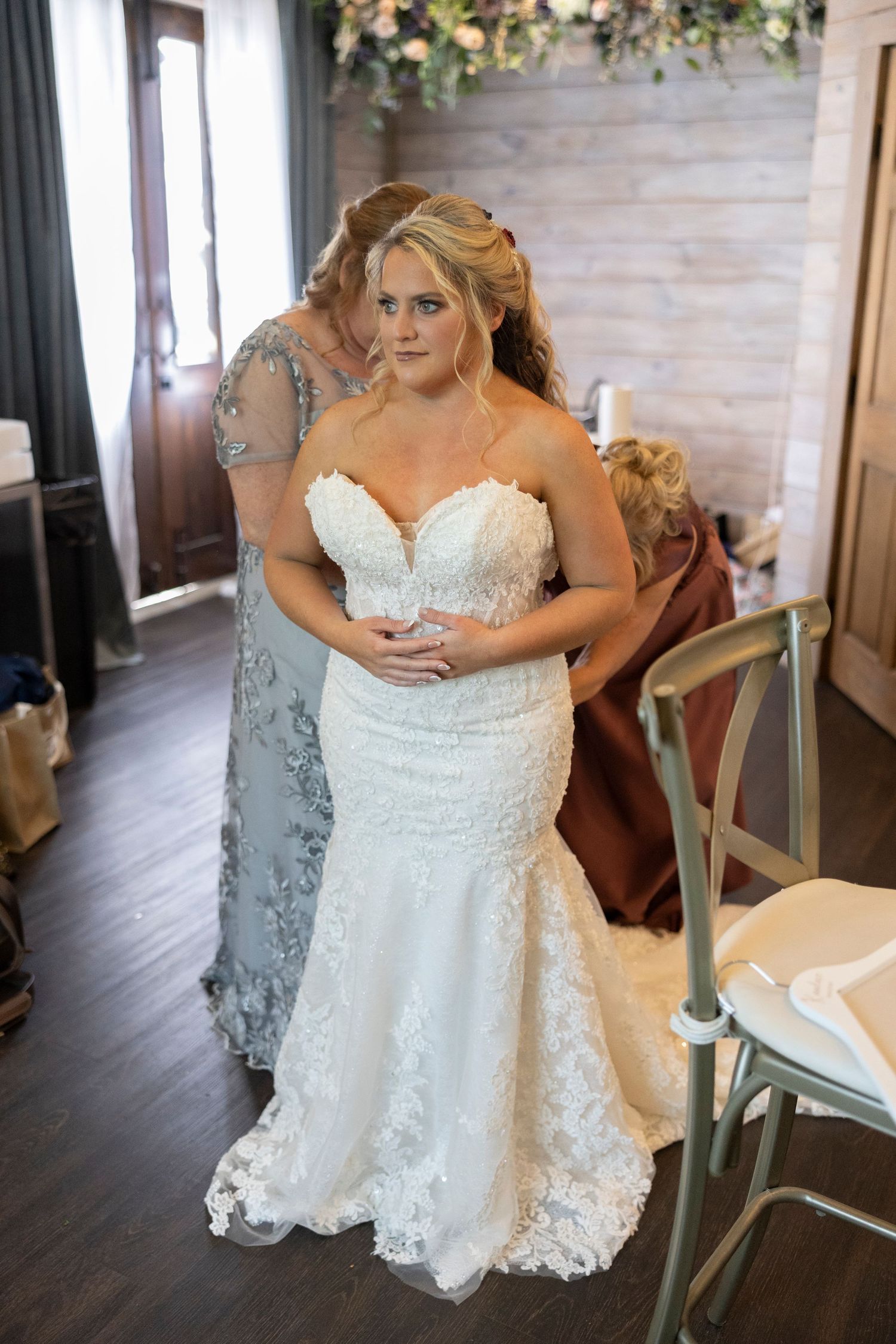 A bride in a white lace wedding dress sits in a rustic venue with wooden floors and natural lighting.
