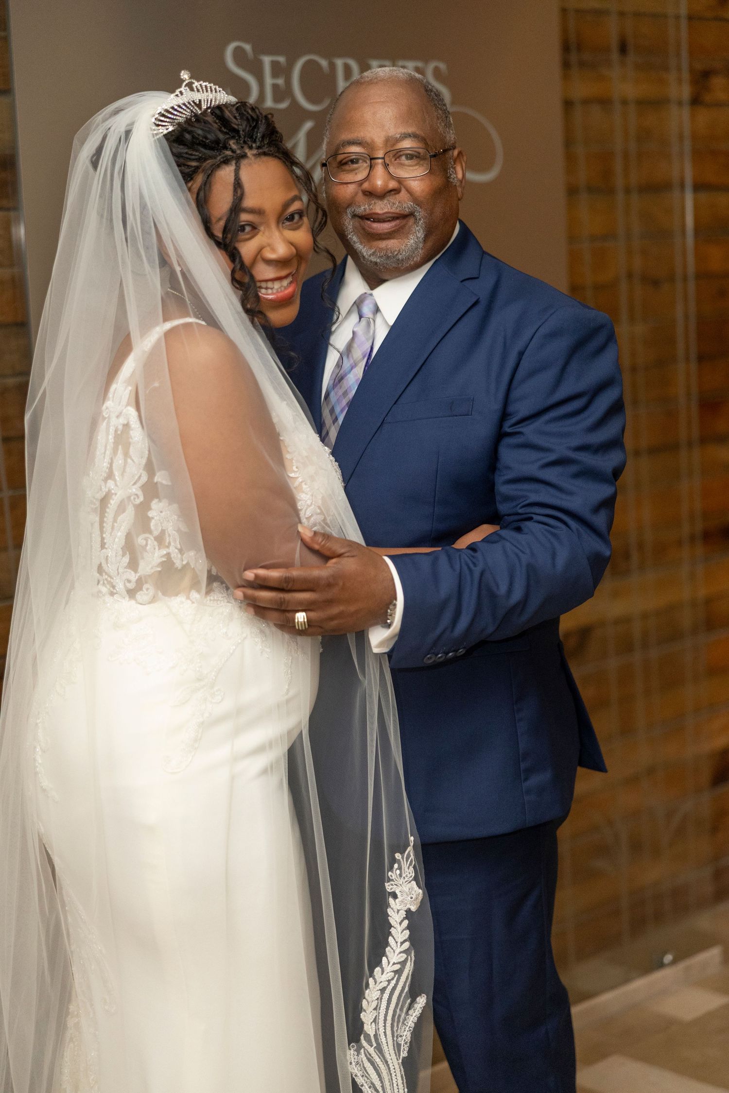 A joyful wedding moment captured showing a bride in a white lace gown with veil embracing someone in a navy suit.