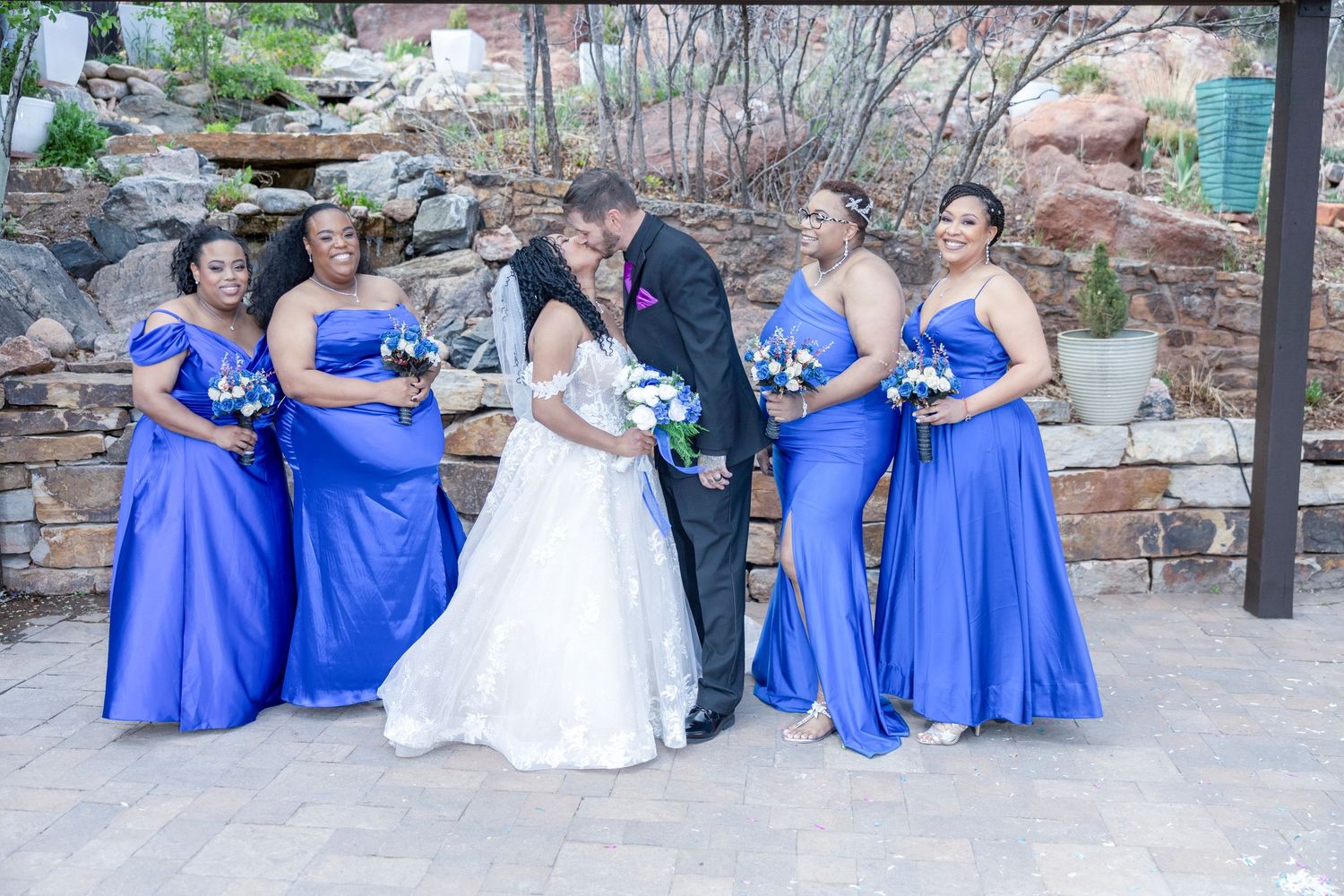 Wedding party in royal blue bridesmaid dresses poses together in an outdoor stone courtyard setting.