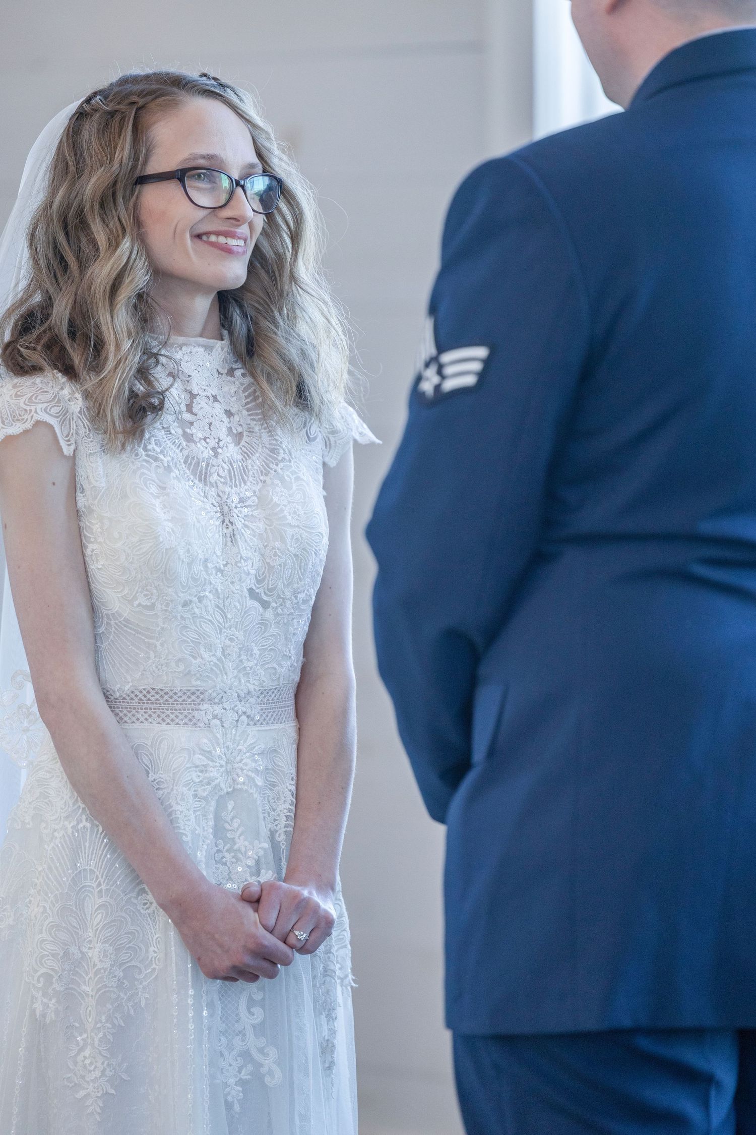 A couple stands together during a wedding ceremony with the bride wearing a white lace dress and groom in navy suit.