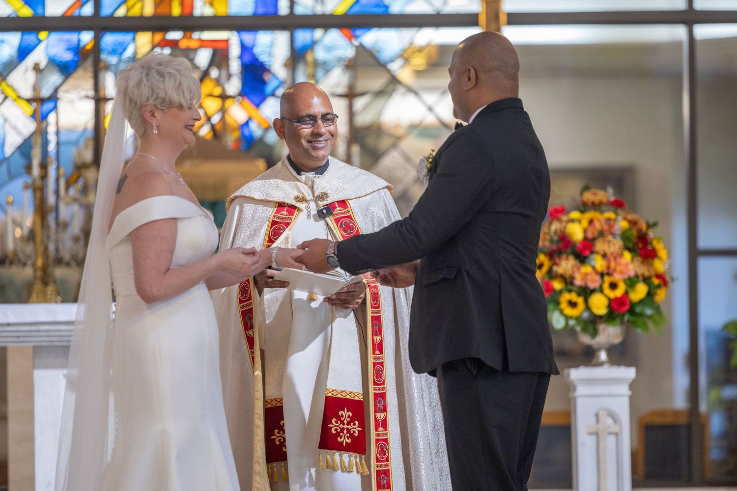 A couple exchanges rings during a traditional church wedding ceremony with colorful stained glass windows in the background.