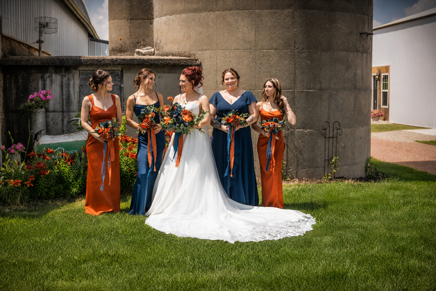 Bride in white gown with bridesmaids in orange and navy dresses holding colorful bouquets outdoors near a rustic silo.