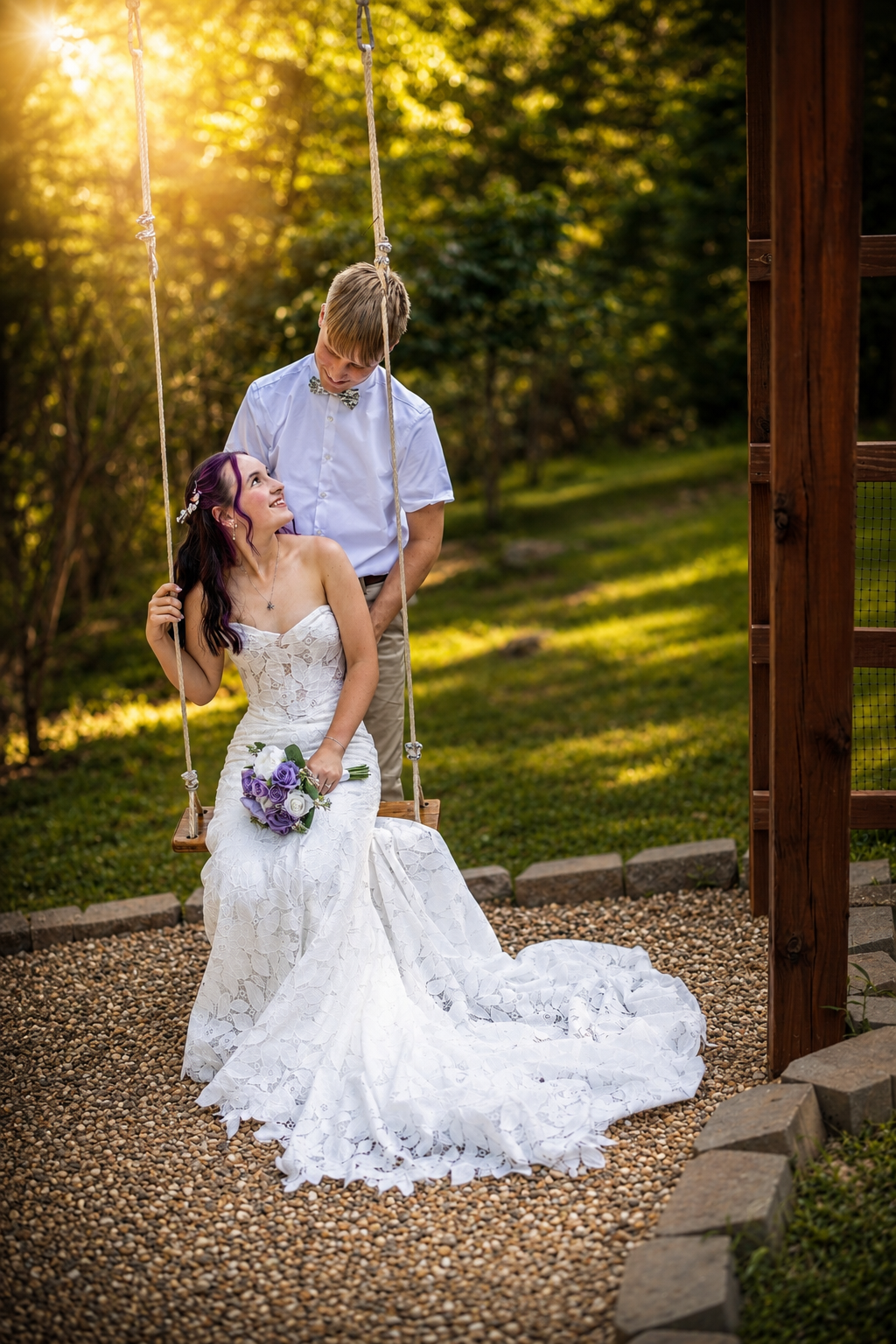 Bride in lace gown sits on garden swing holding purple bouquet as groom stands behind her in golden sunset light.