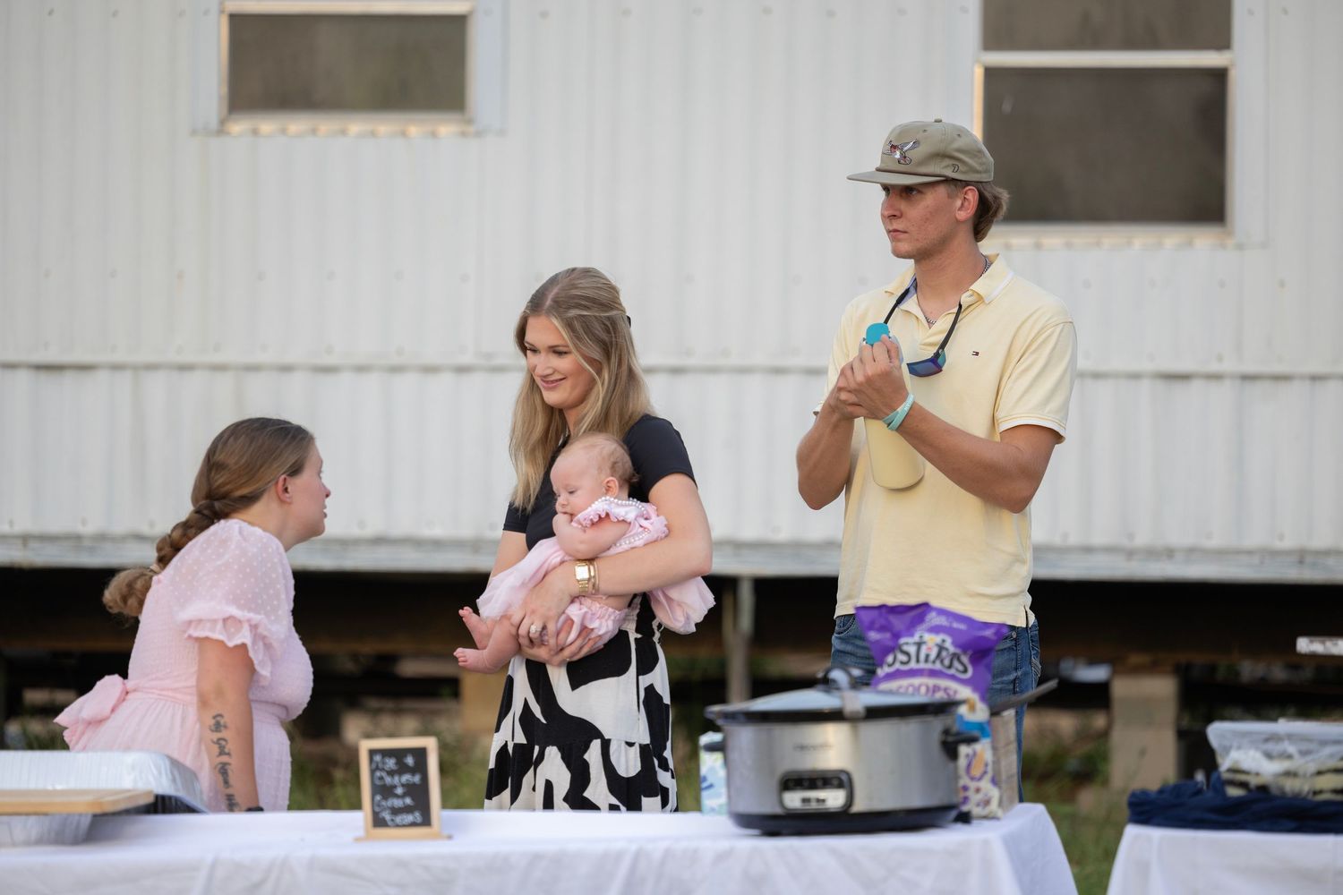 Family gathering outdoors with woman holding baby, young girl, and man standing near a table with a slow cooker.