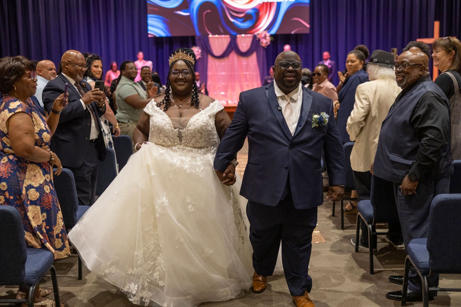Joyful newlyweds walk down the aisle at their wedding ceremony, surrounded by smiling guests in a decorated venue.