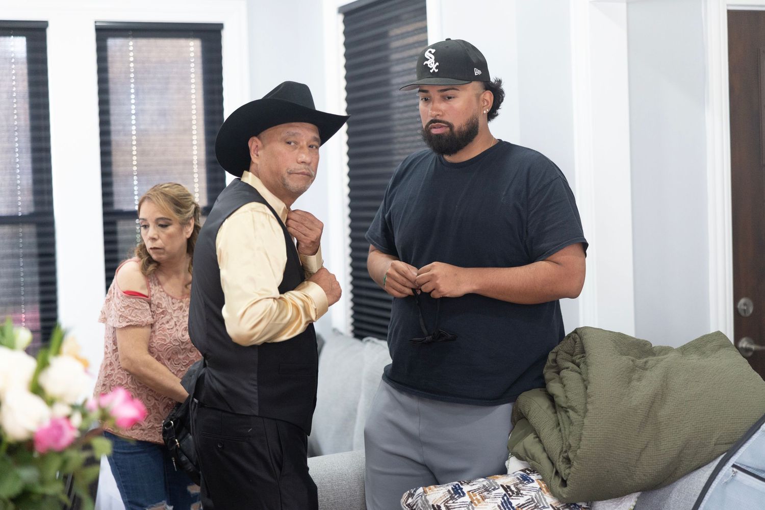 Three people stand indoors, one man wearing a cowboy hat and vest, another in a black t-shirt, and a woman nearby.