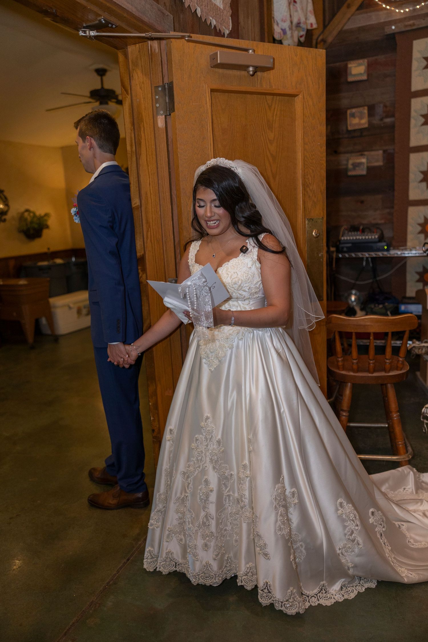 Bride in white ball gown reads vows while holding groom's hand during indoor rustic barn wedding ceremony.