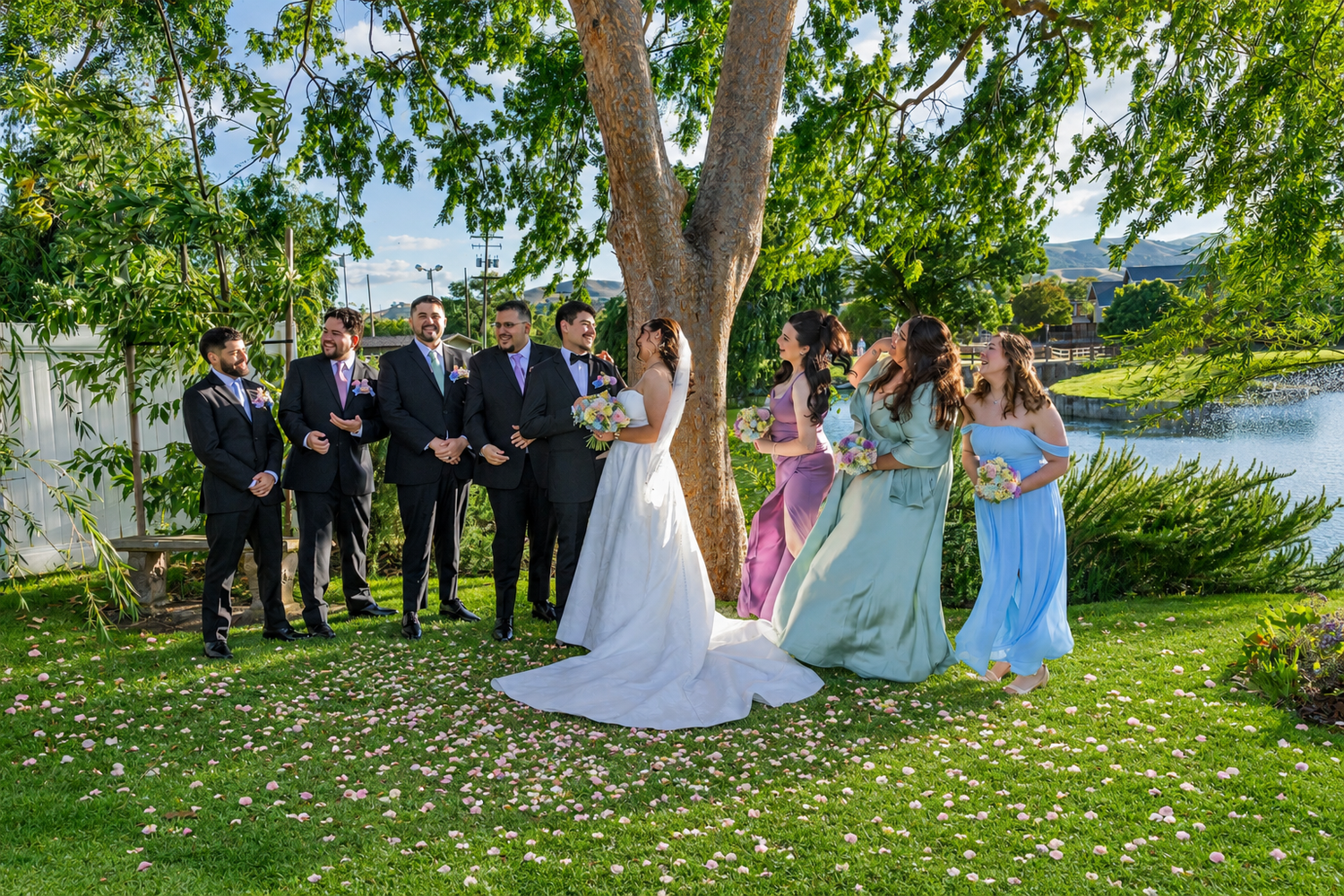 Wedding party poses outdoors under a large tree by a lake, bride in white gown with groomsmen and bridesmaids in blue.