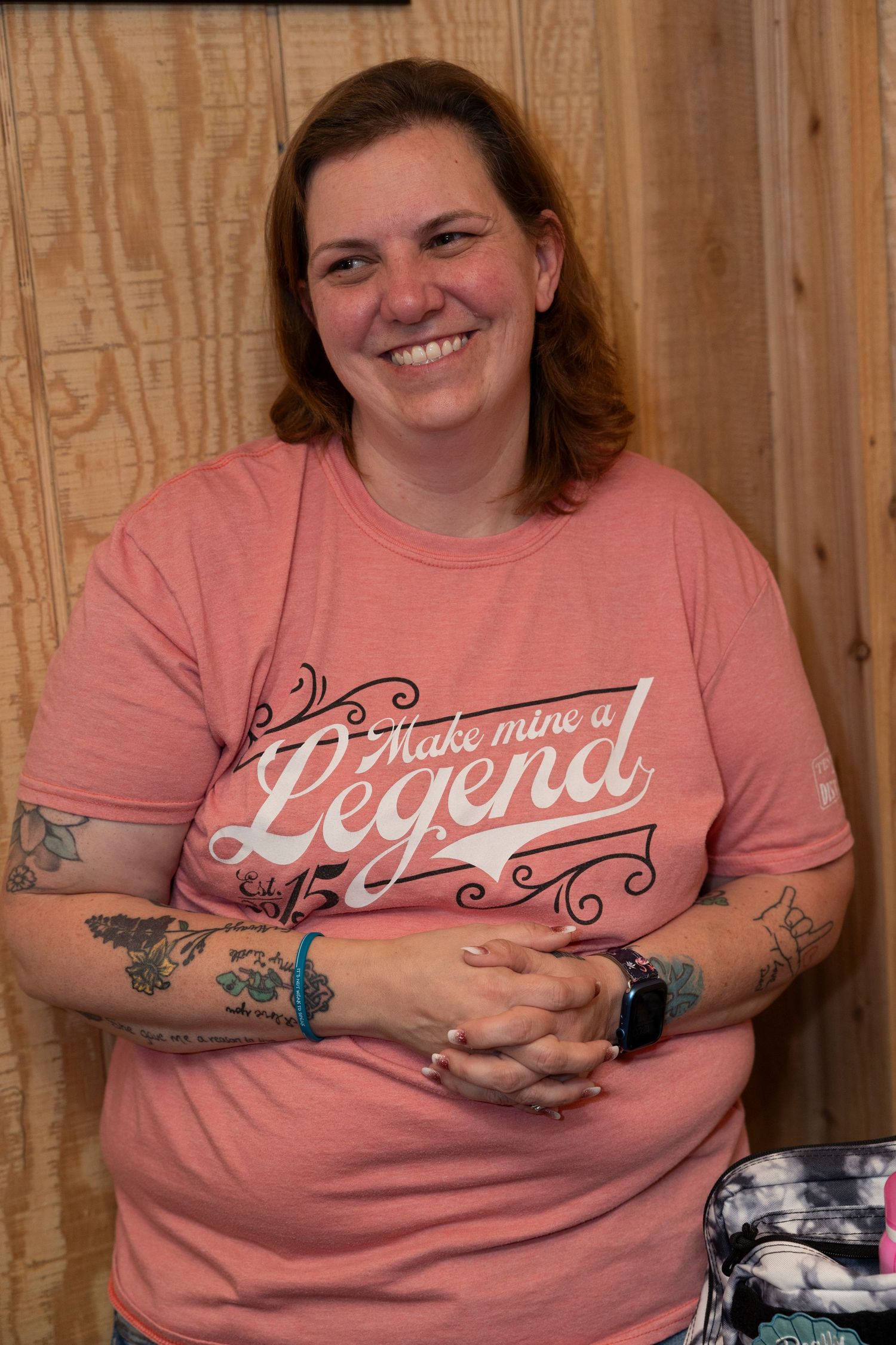 A person in a pink t-shirt with decorative text design smiles while wearing visible arm tattoos.