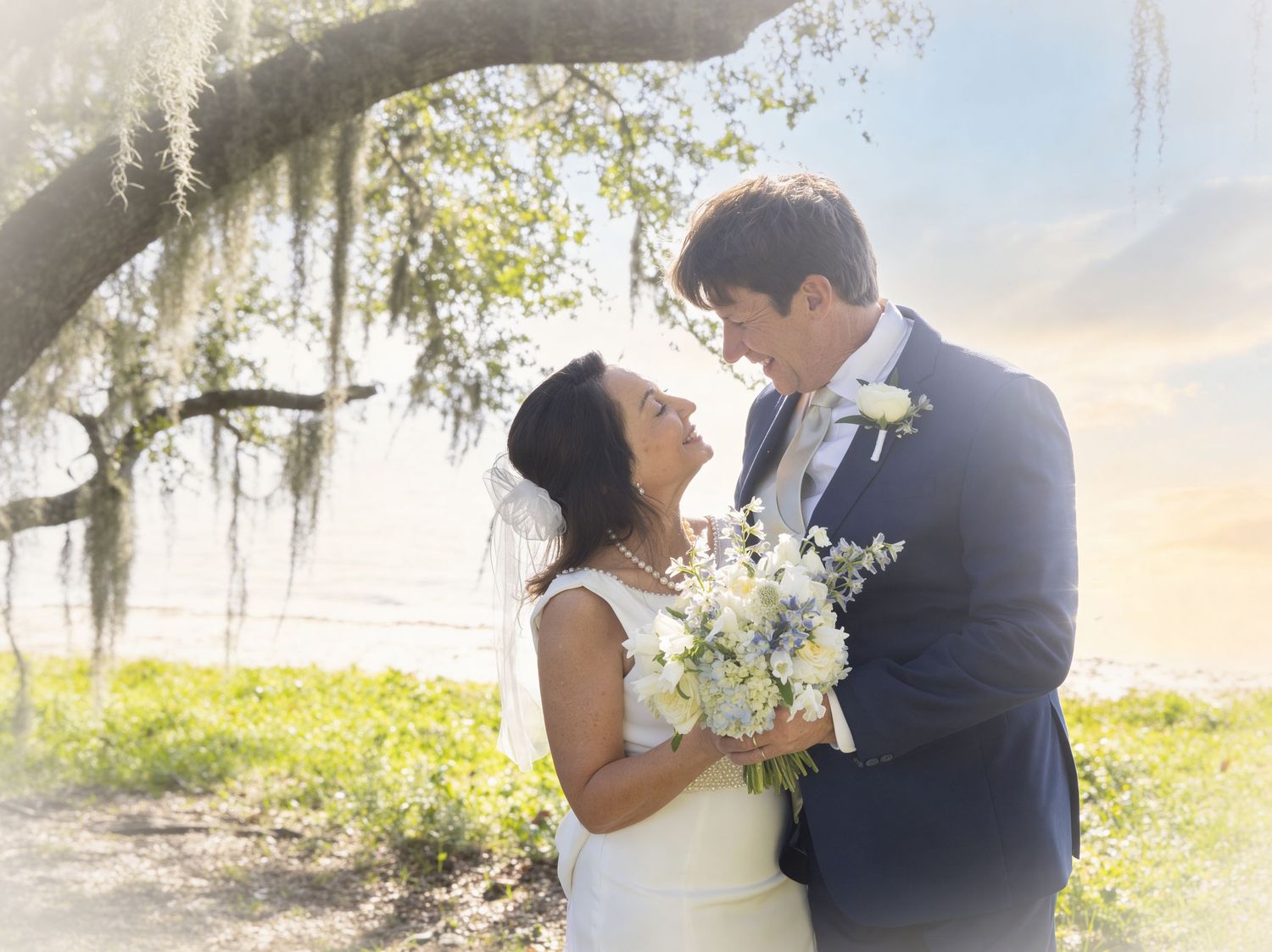 Bride and groom share a romantic moment under a moss-draped oak tree near the water on their wedding day.