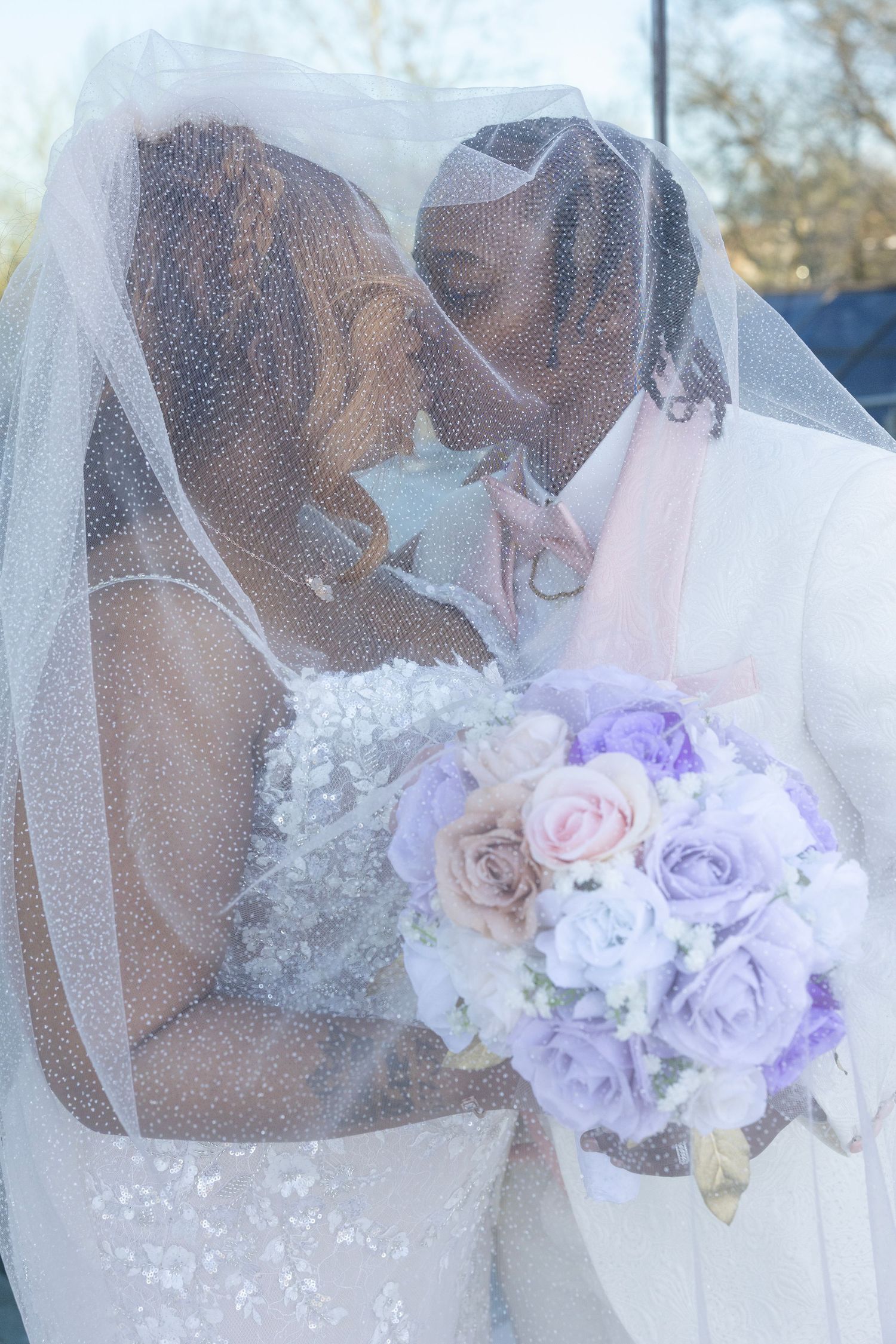 A Black bride and groom share a romantic kiss under a white veil, bride holding a purple and pink rose bouquet.