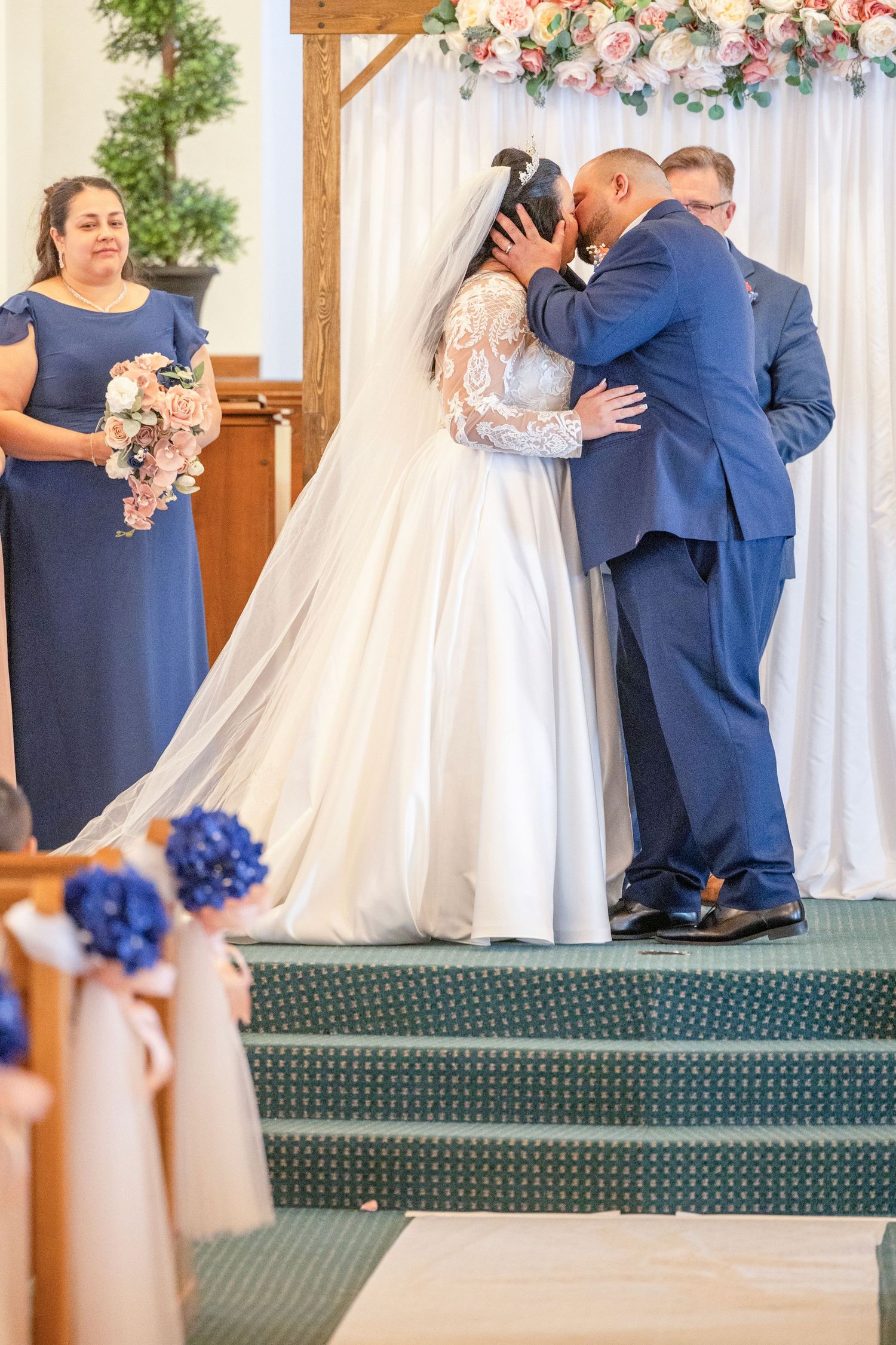 Bride and groom share first kiss at church altar, bridesmaid in blue dress holding bouquet stands nearby.