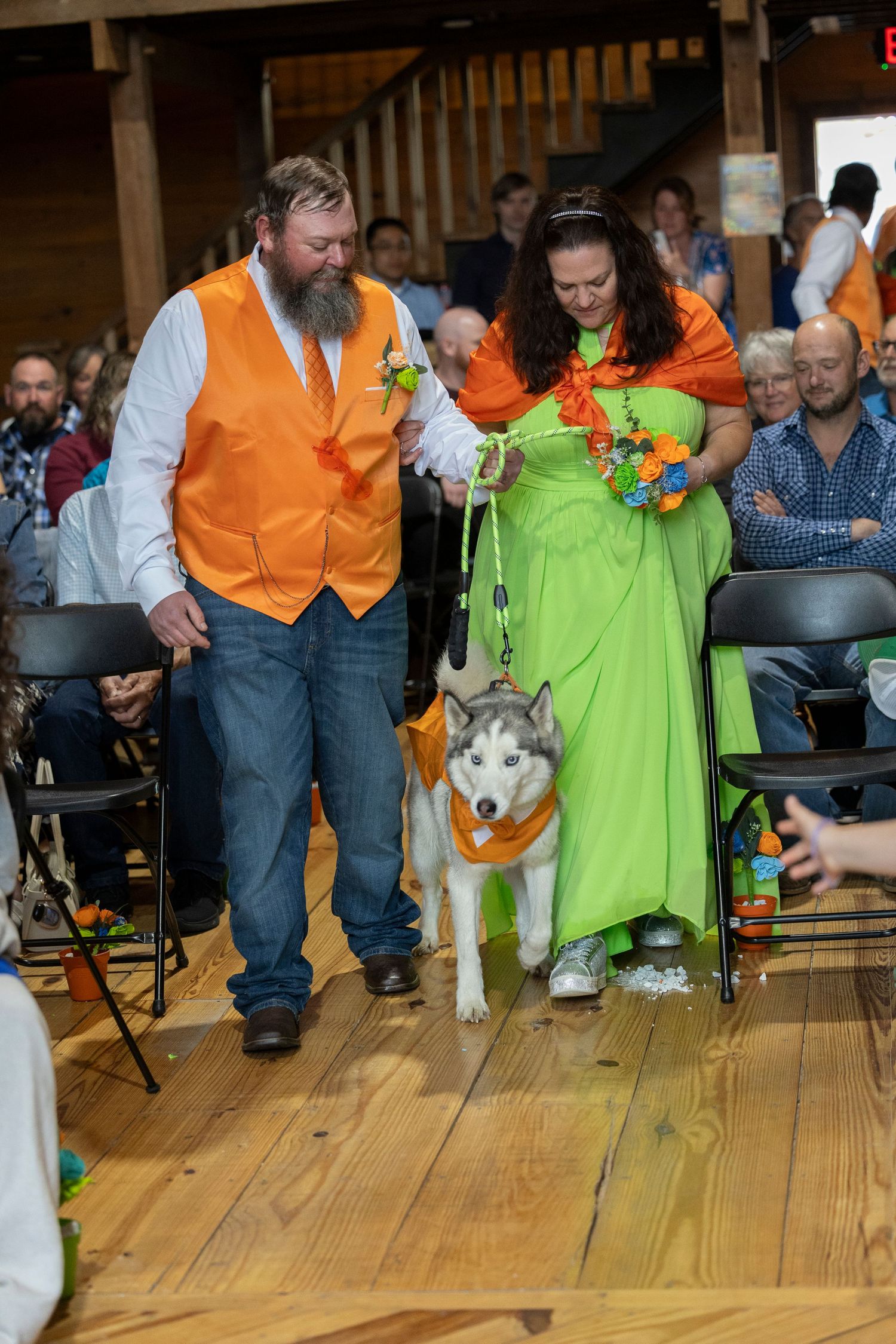 Couple in orange and green attire walk a Husky dog down the aisle at a colorful indoor wedding ceremony.