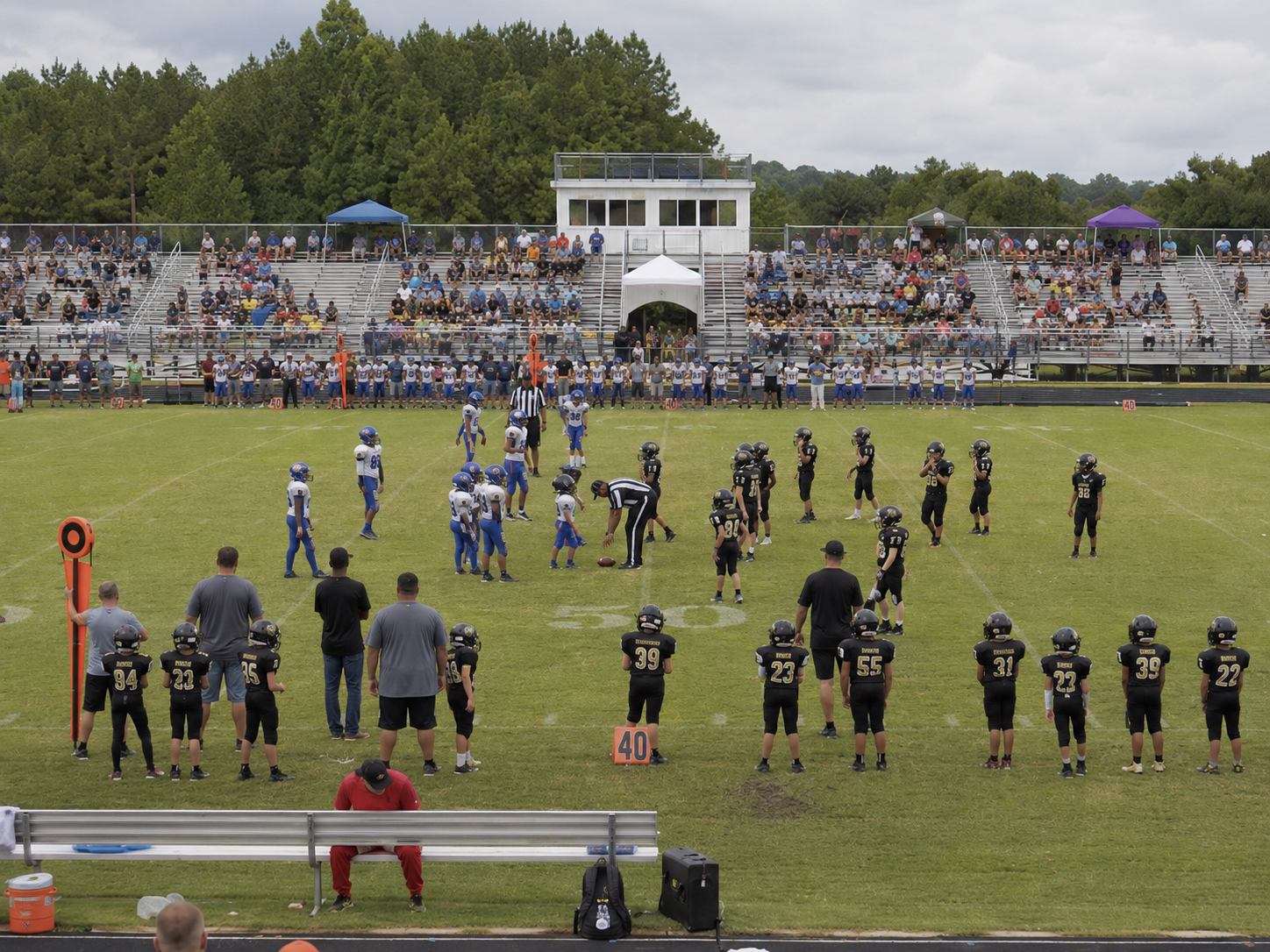 Youth football teams face off on a green field stadium with packed bleachers of spectators on a sunny day.