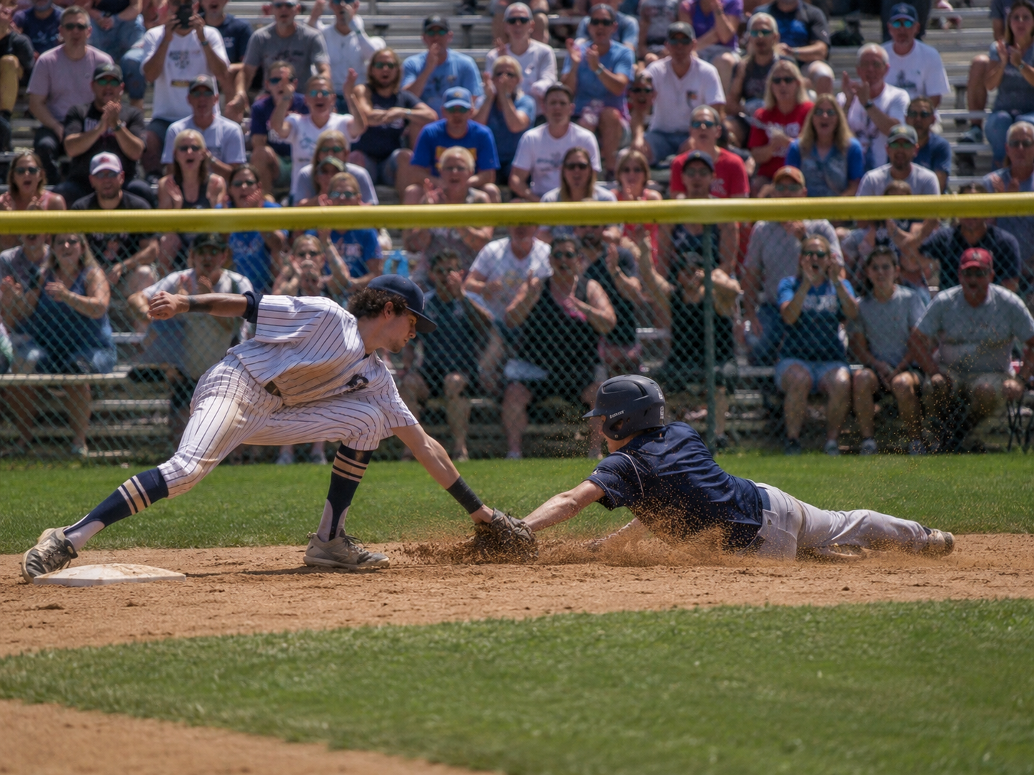Baseball player slides into base as fielder attempts a tag play during a crowded outdoor game.