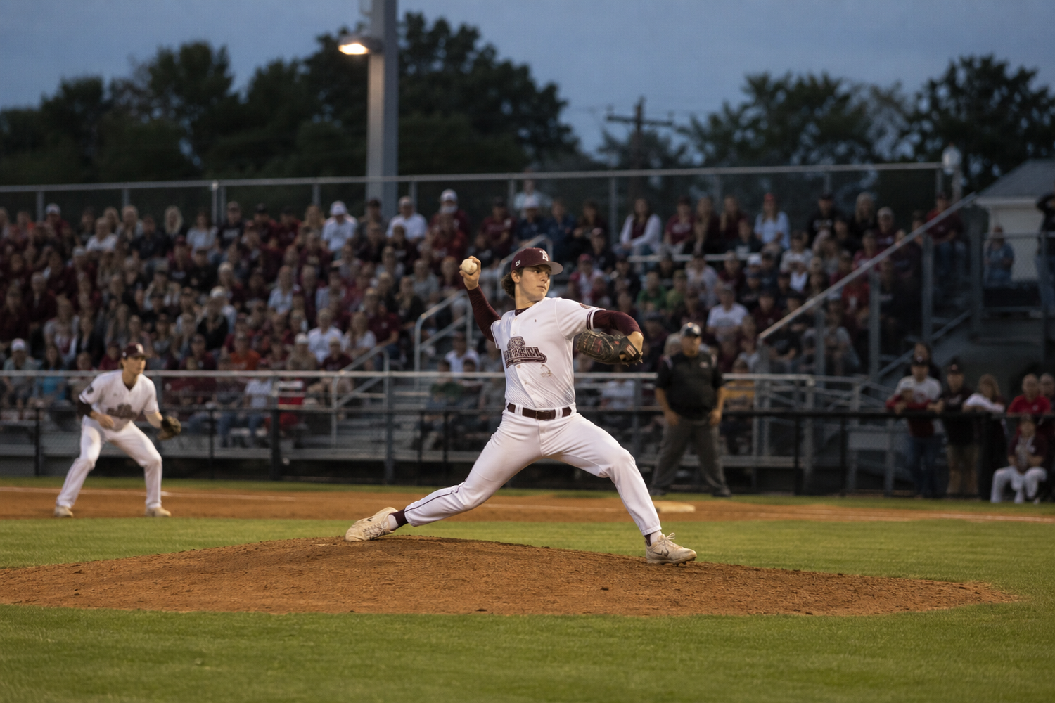 Baseball pitcher in white uniform winds up to deliver a pitch during a night game before a full crowd.