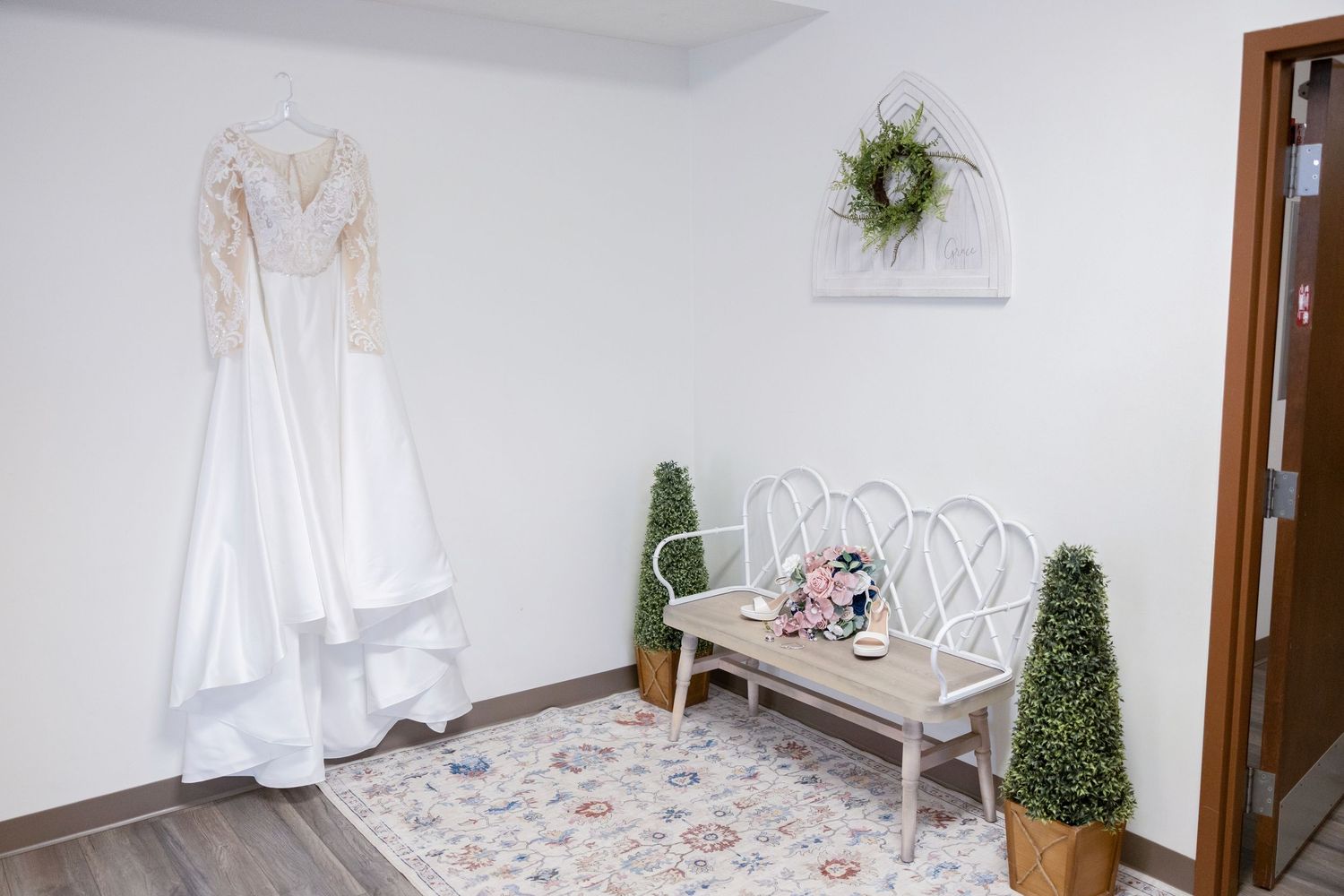 Elegant bridal suite with a white wedding gown hanging on wall, vintage bench, floral bouquet, and topiary plants on a patterned rug.