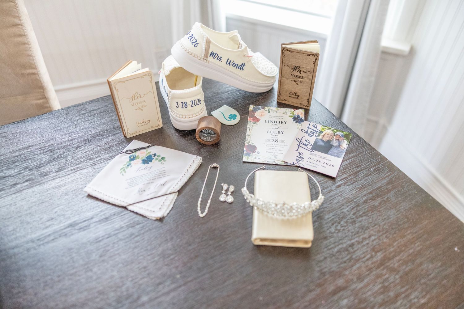 Wedding flat lay with bridal accessories, tiara, shoes, invitations, and jewelry on a dark wood table.
