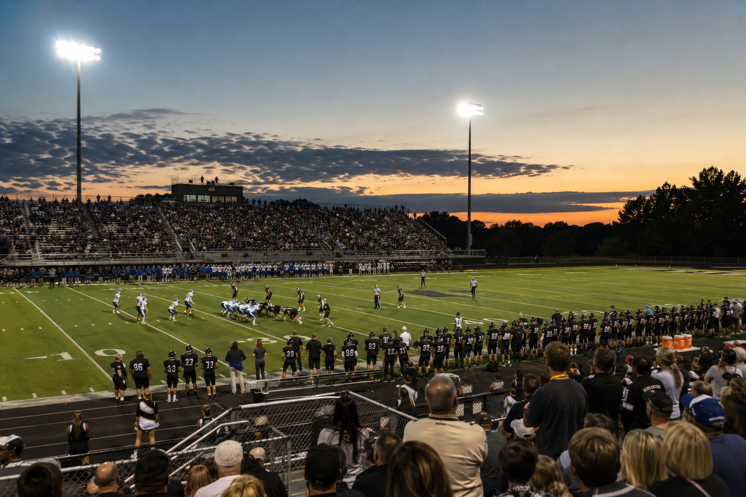 Aerial view of a high school football game at dusk with stadium lights and colorful sunset sky over packed bleachers.