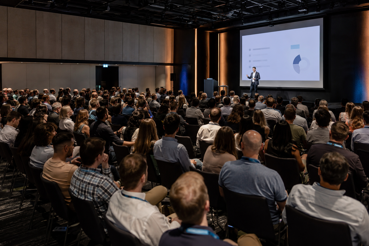Large audience attending a professional conference presentation with a speaker on stage and a projected screen.