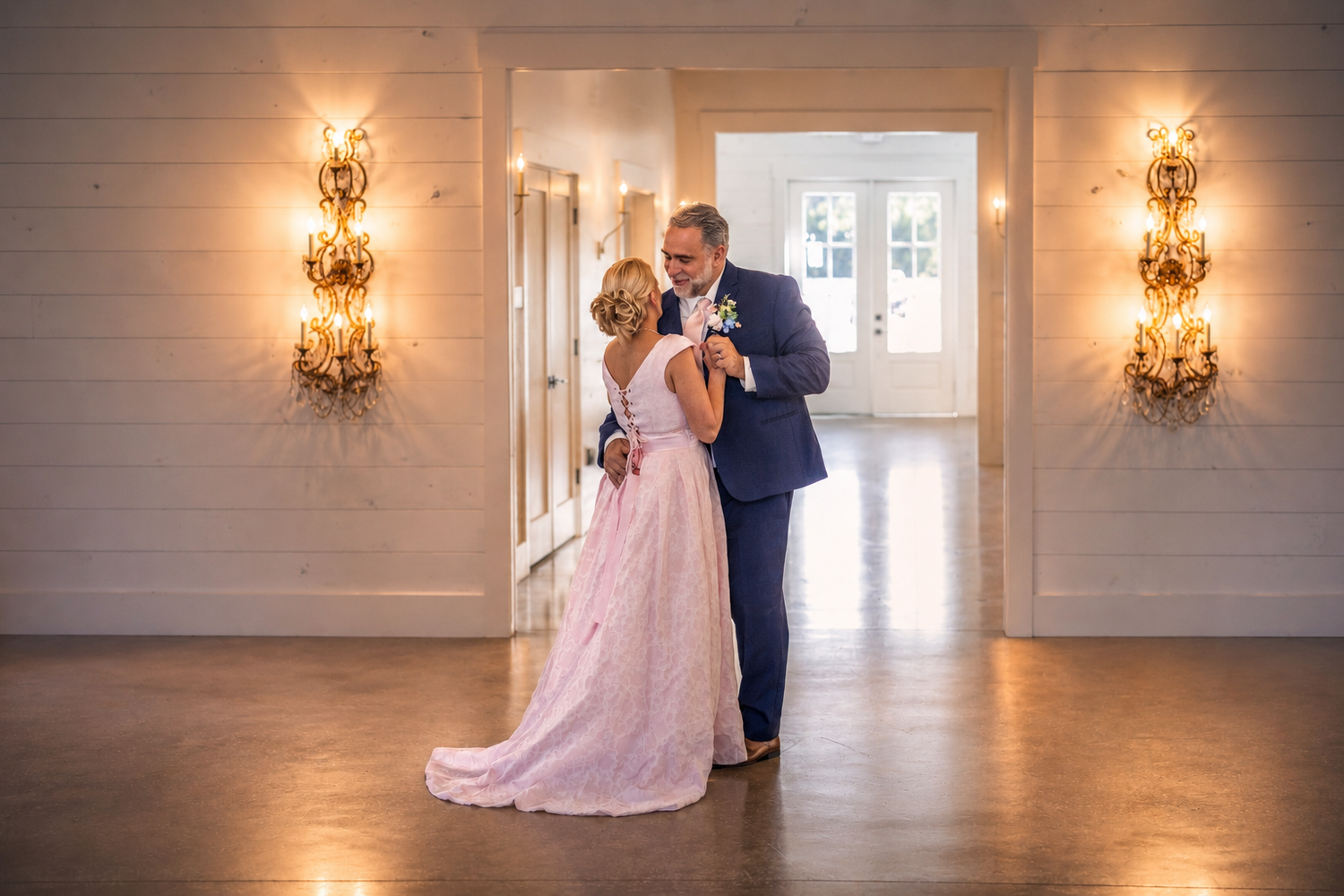 Couple sharing a tender dance in an elegant white hall adorned with golden wall sconces.