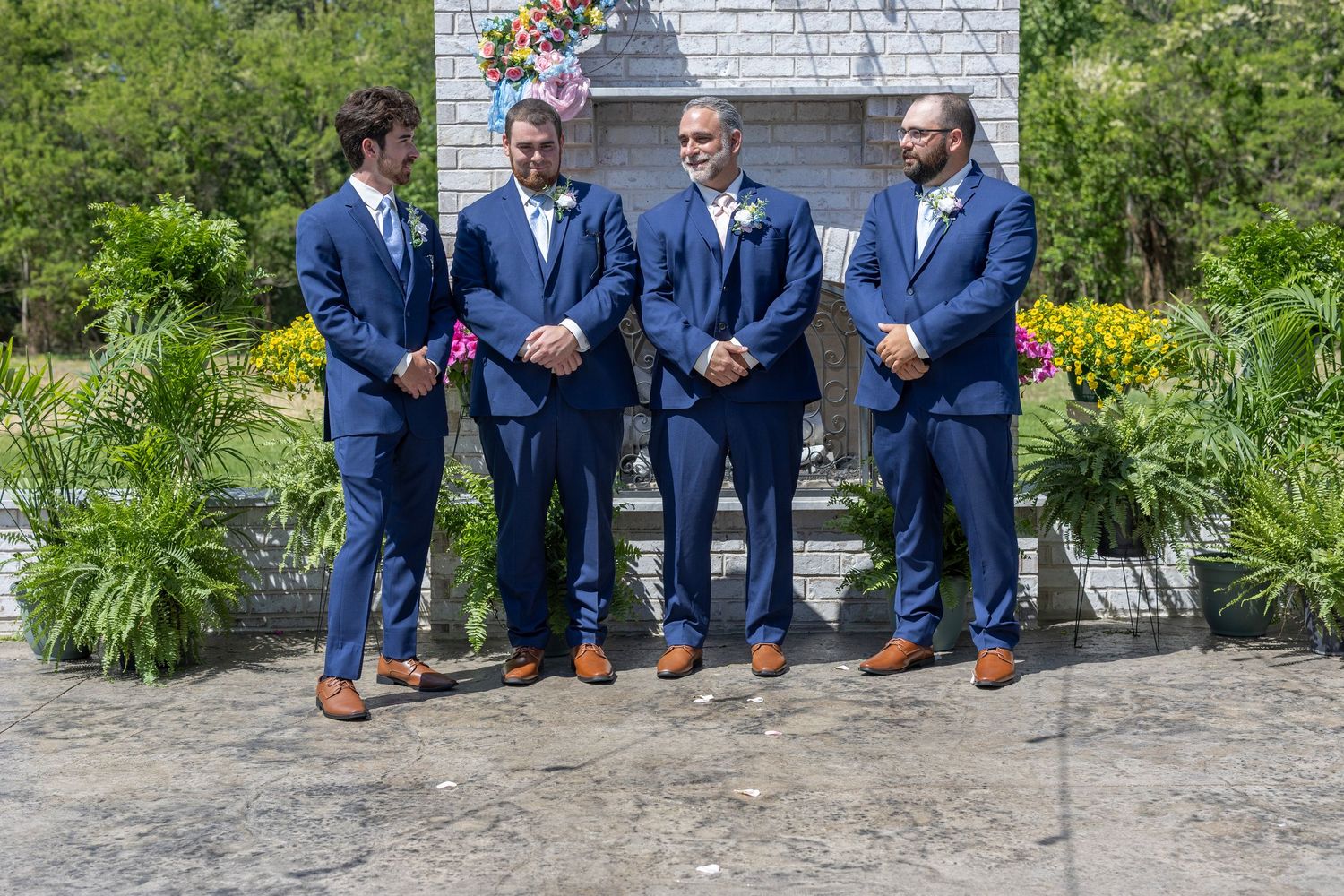 Four groomsmen in navy blue suits with boutonnieres standing outdoors near a stone monument on a sunny day.