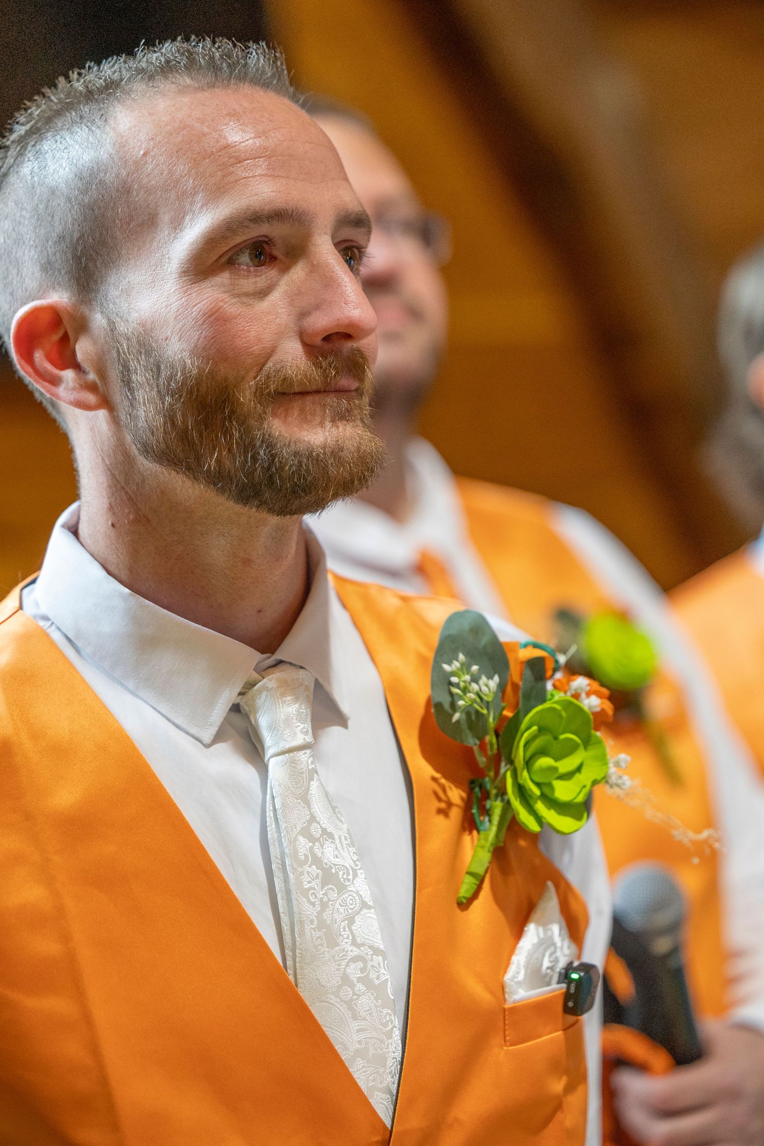 Man in orange suit with floral boutonniere at wedding ceremony, wearing white lace tie, emotional expression.
