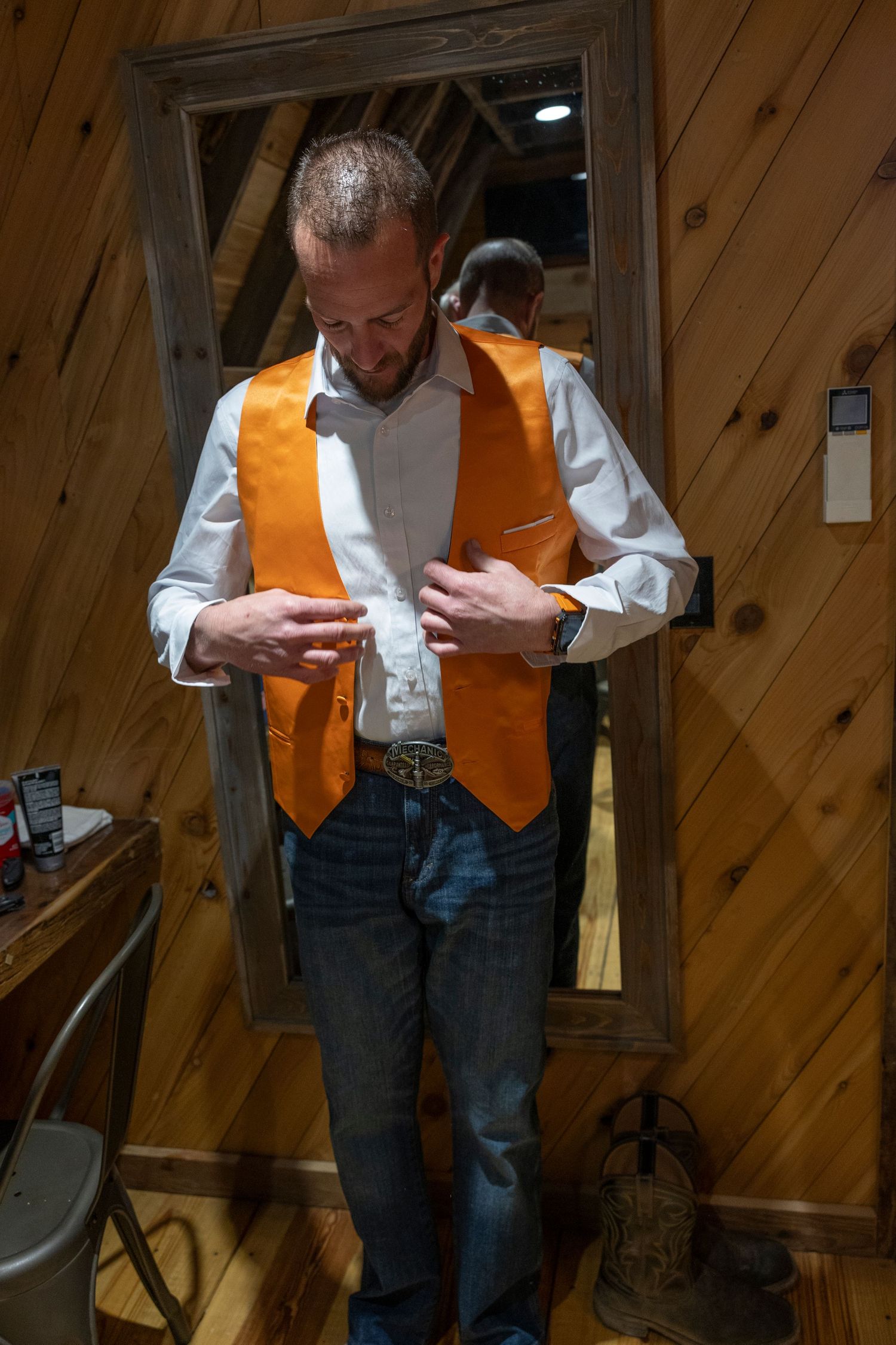 Man in orange vest and jeans buttoning shirt in rustic wood-paneled room with mirror reflection.