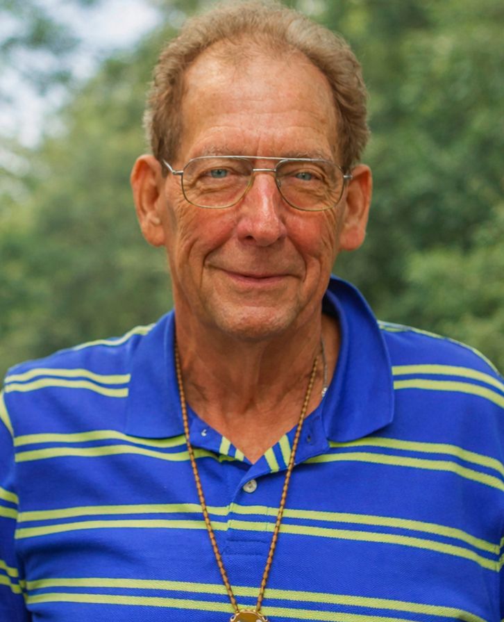 Smiling older man with glasses wearing a blue striped polo shirt and gold necklace outdoors.