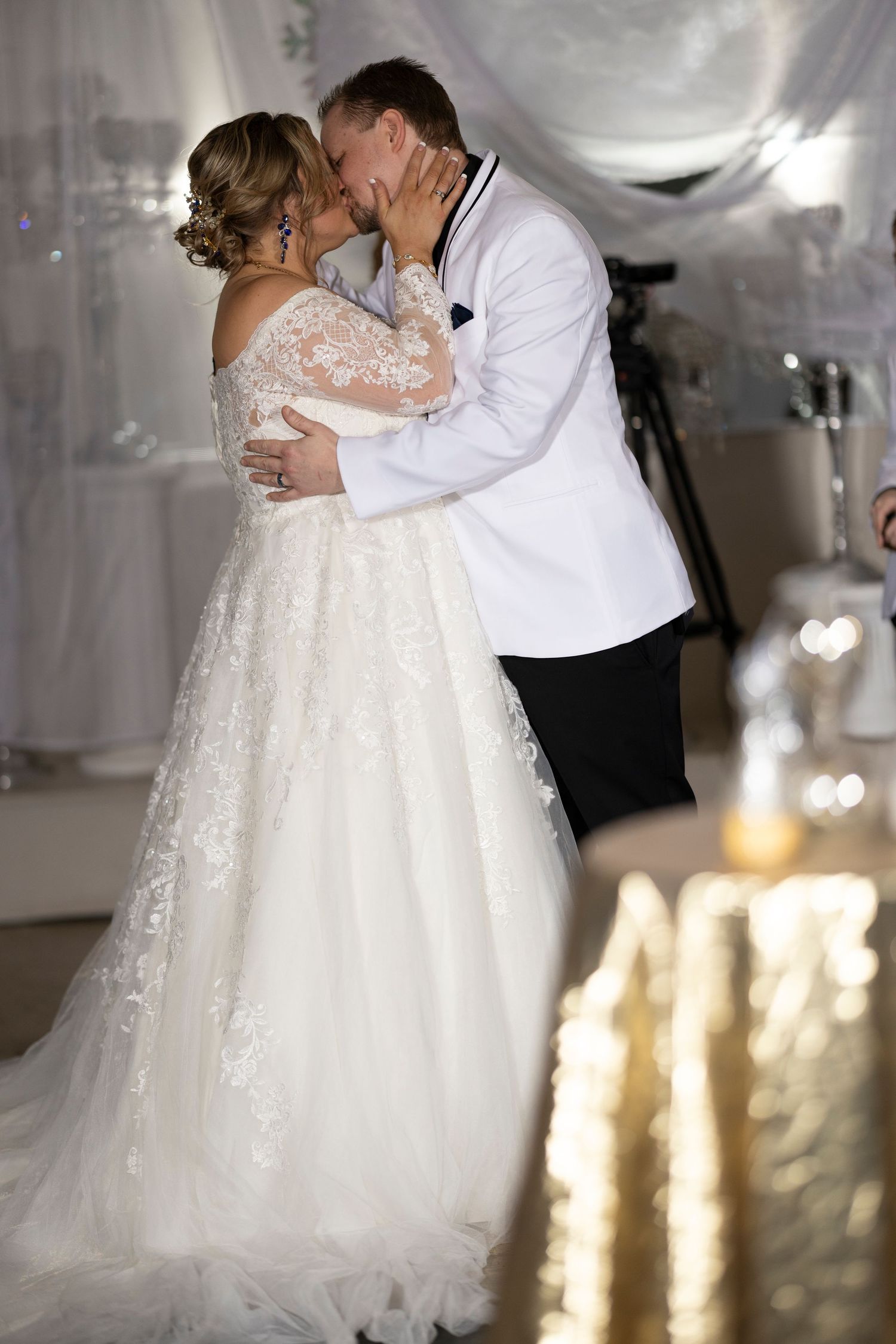 Bride and groom share first dance kiss, bride in lace ballgown, groom in white tuxedo jacket at elegant wedding reception.