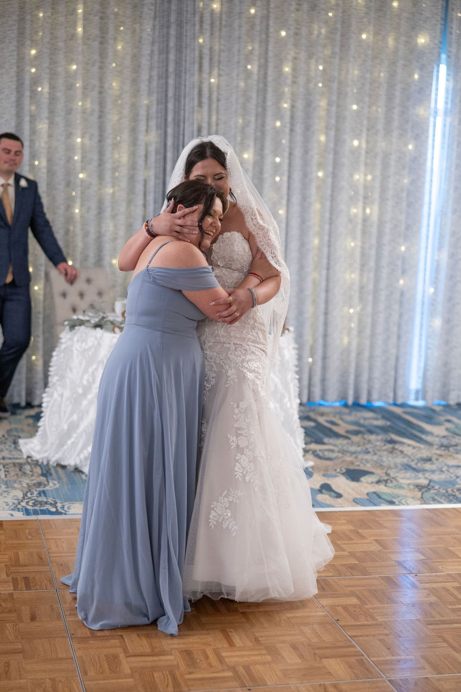 Bride in lace gown hugging bridesmaid in blue dress on dance floor at elegant wedding reception.