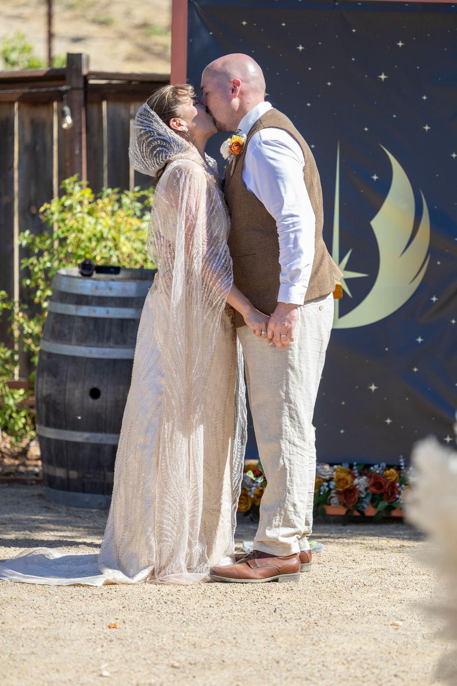 Bride and groom share first kiss at outdoor rustic wedding ceremony with moon backdrop and wine barrel.