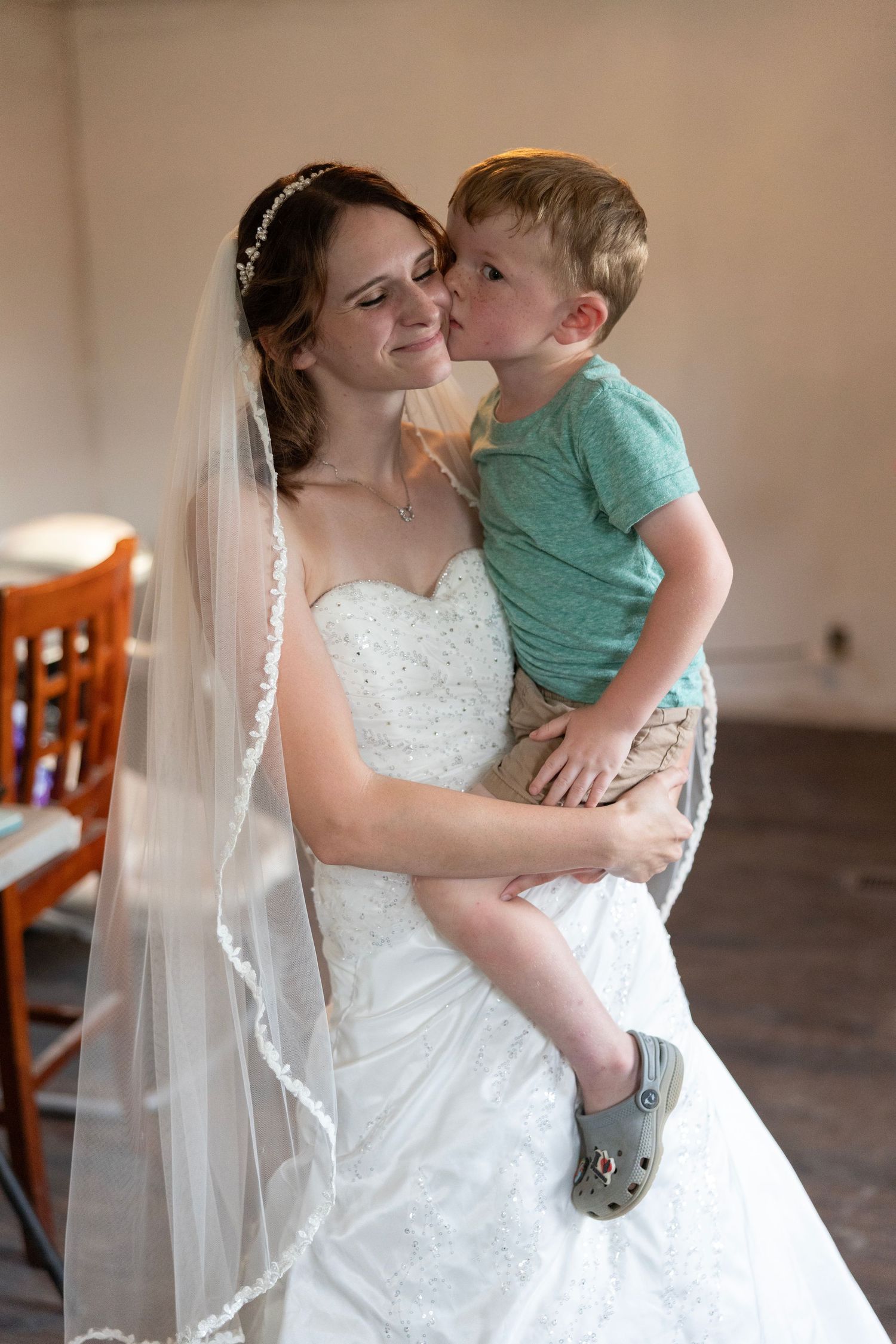 Bride in white gown and veil holds young boy who kisses her cheek during wedding day celebration.