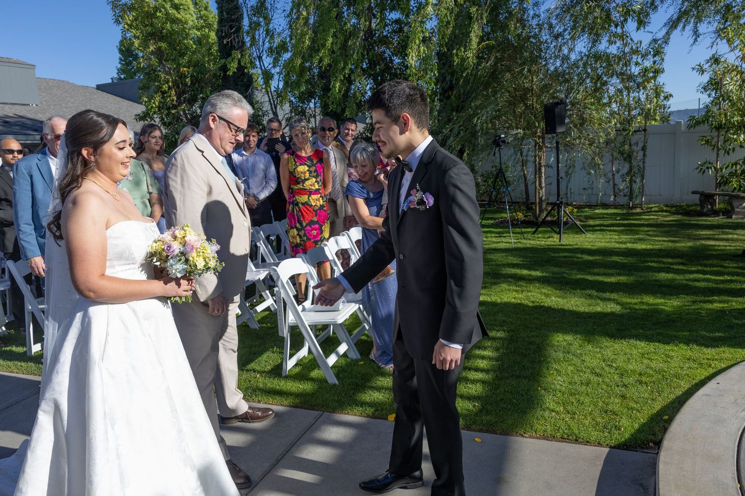 Bride and groom exchanging vows outdoors at a sunny garden wedding ceremony with guests seated in white chairs.