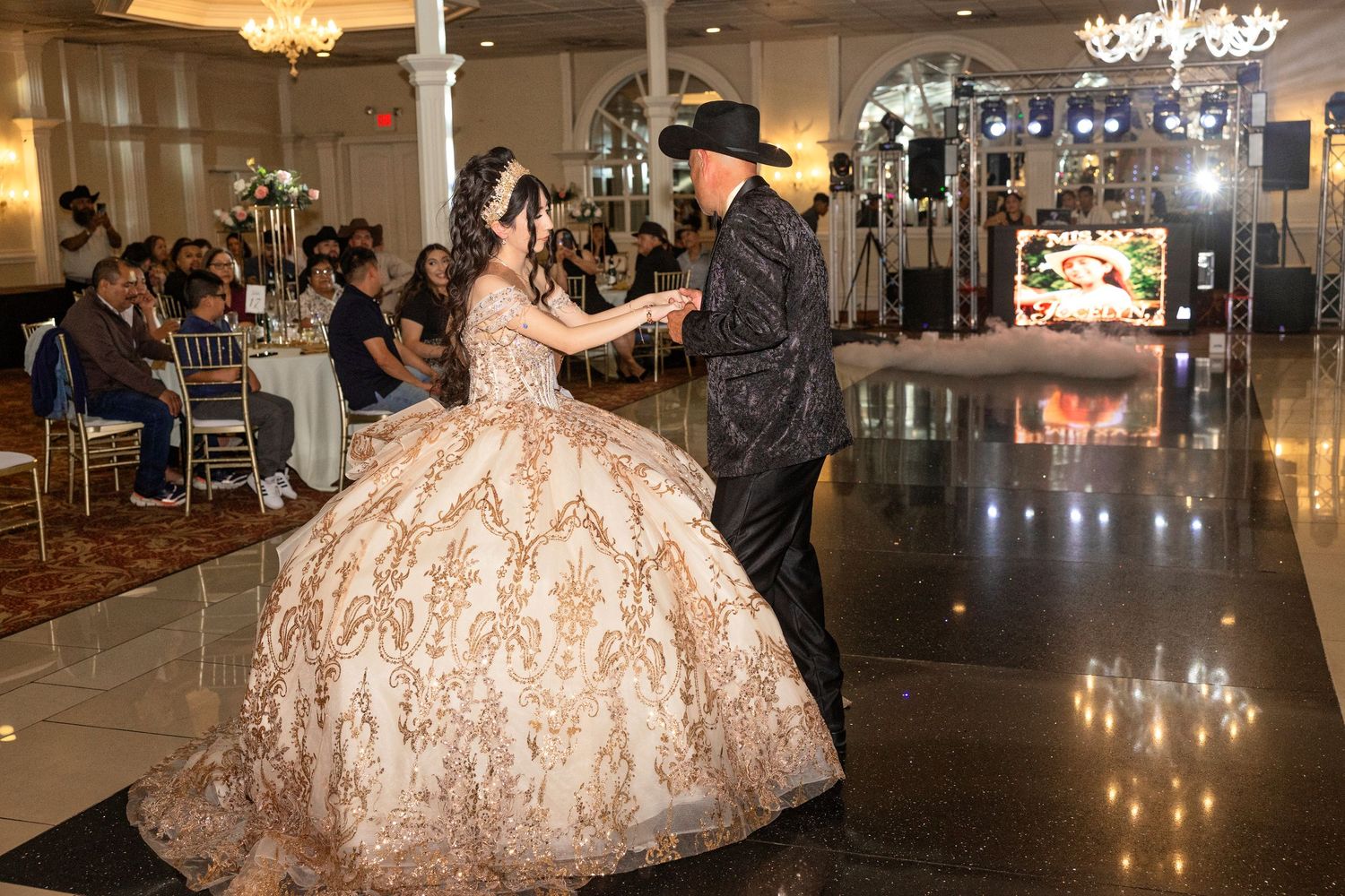 A young woman in a gold ballgown dances with a man in a black suit at an elegant quinceañera celebration.