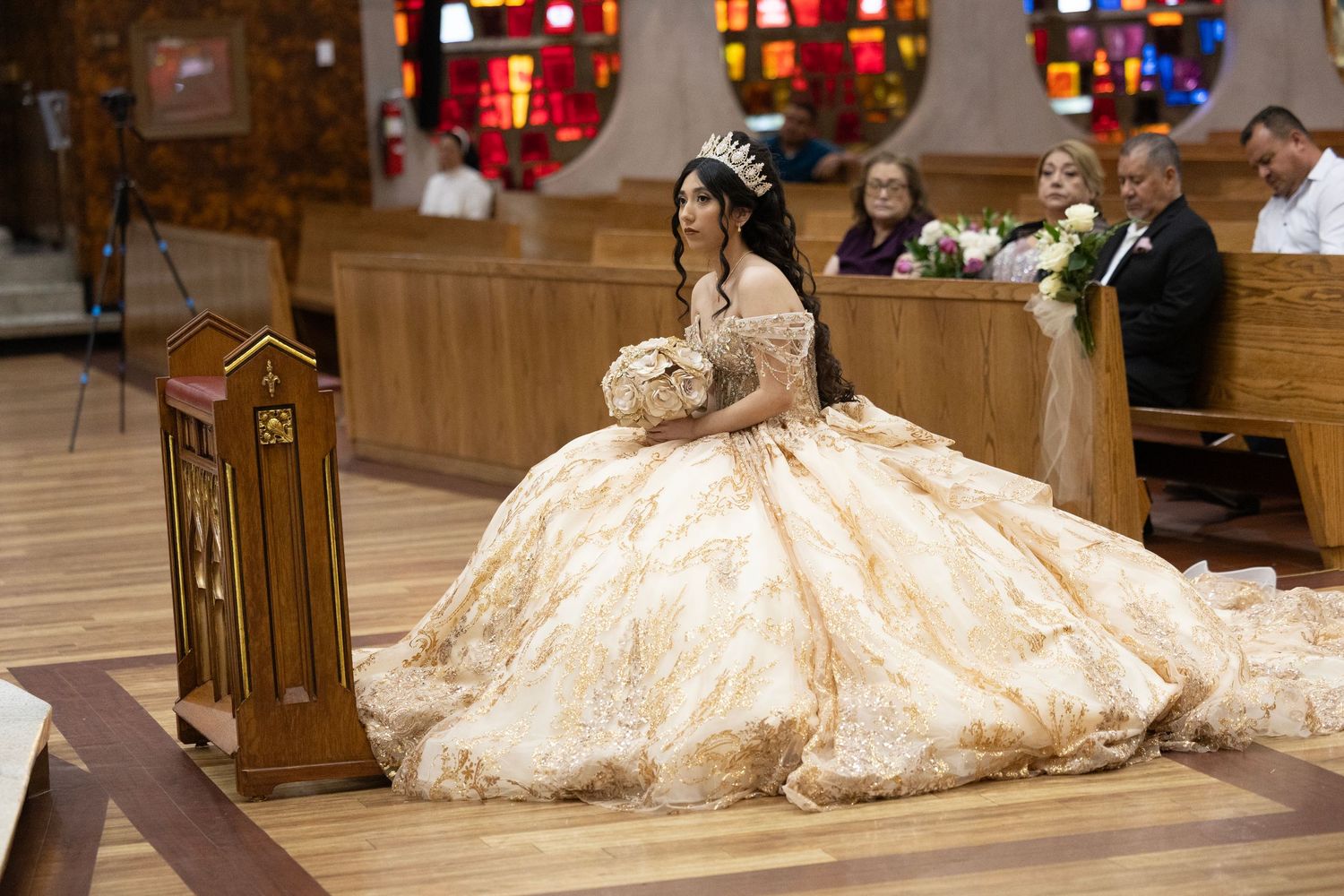 Young woman in elegant gold quinceañera gown kneeling at church pew during ceremonial celebration.